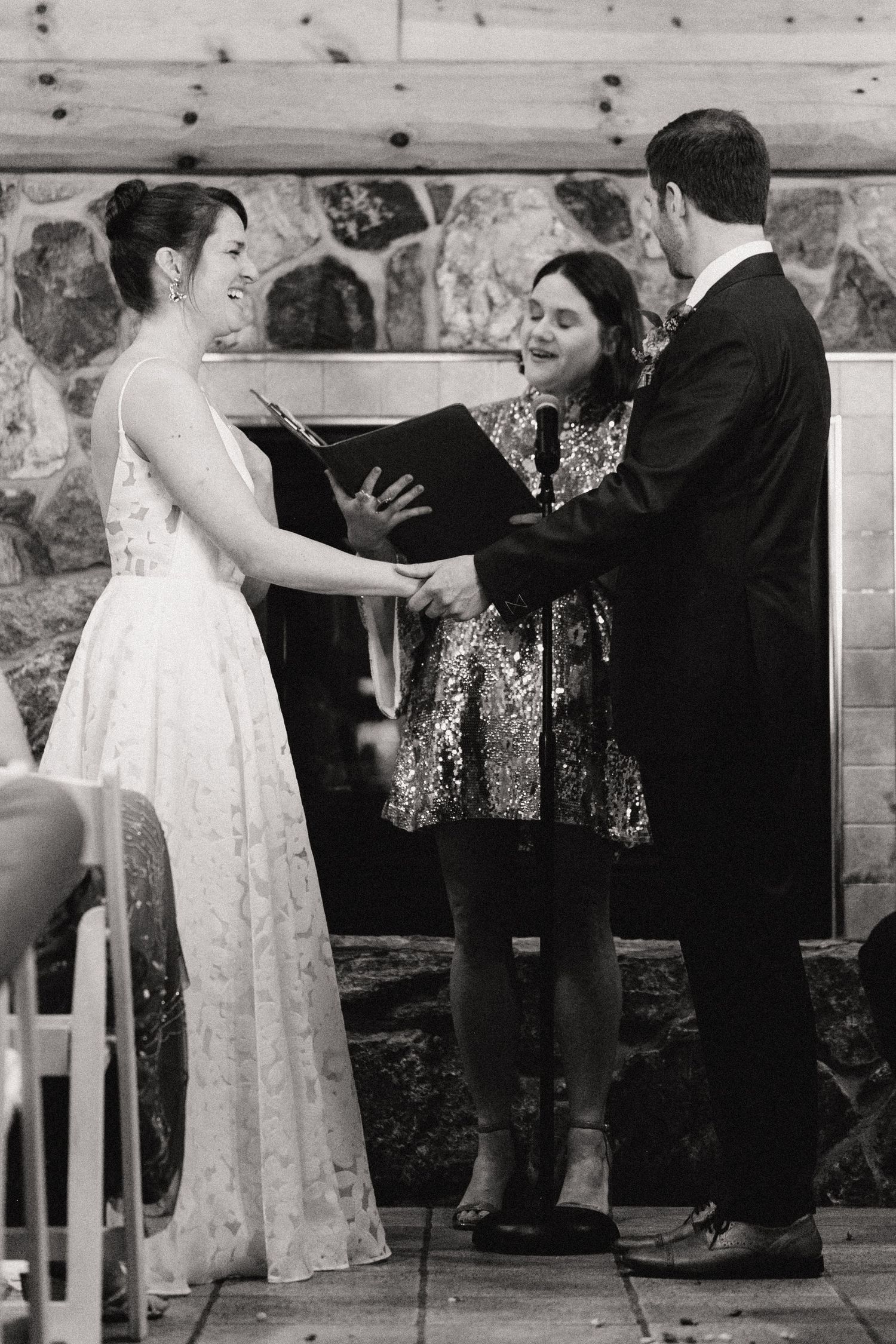 A wedding ceremony takes place in front of a rustic stone wall while an officiant in patterned dress presides over the service.