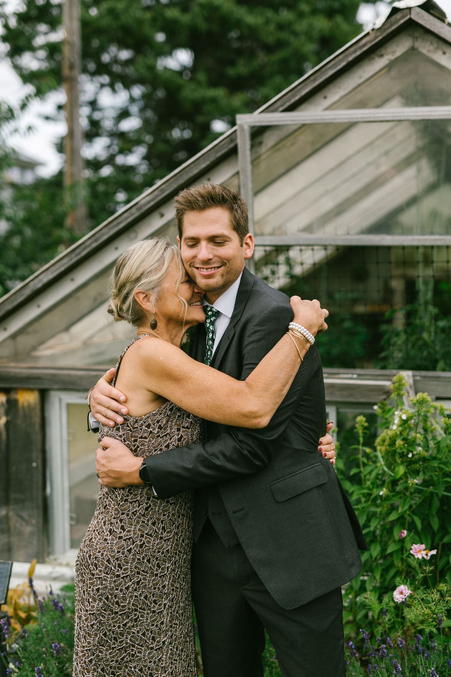A mother and son embrace and laugh together in front of a greenhouse with lush greenery in the background.