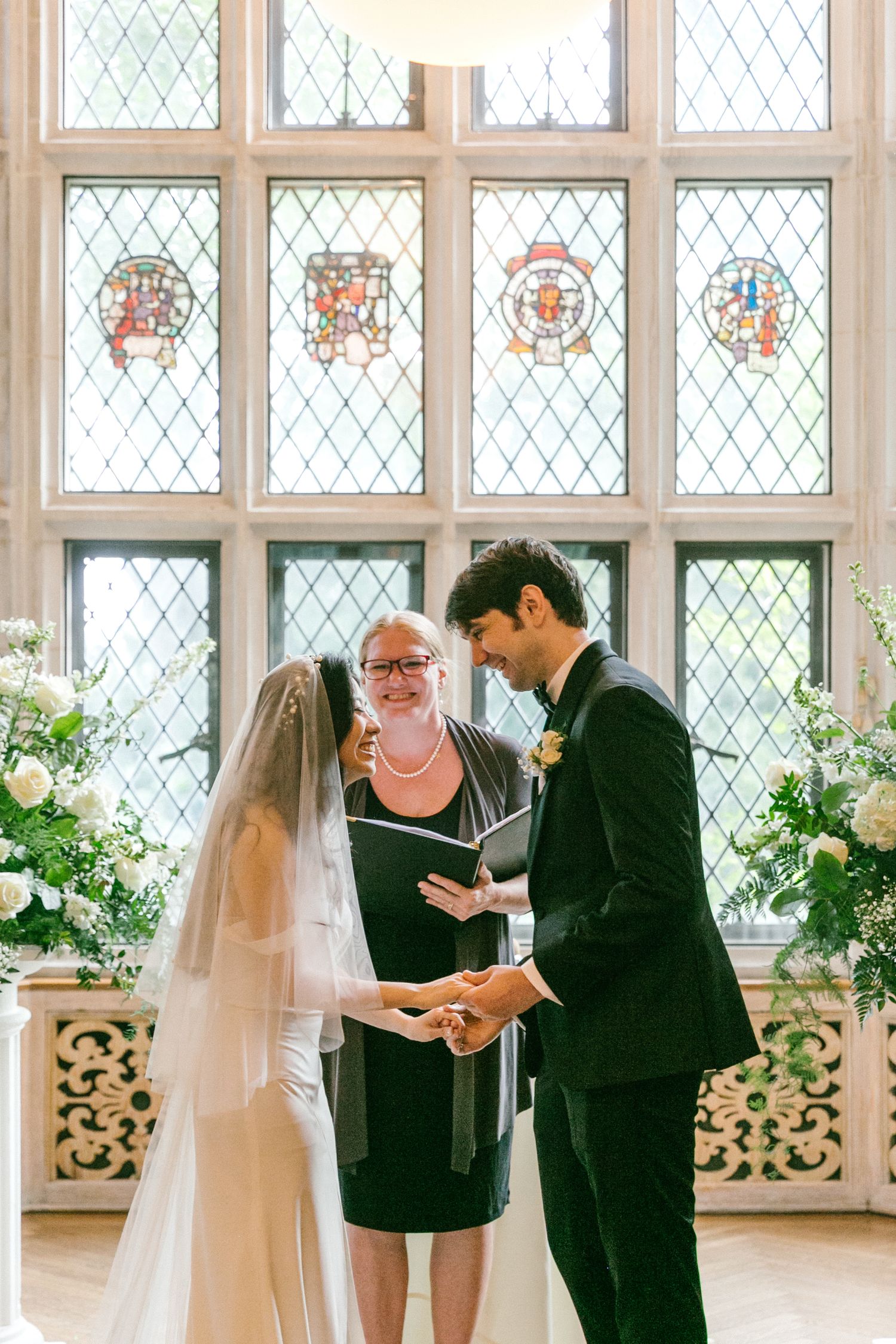 Bride and groom exchange vows before an officiant with stained glass windows and white floral arrangements.