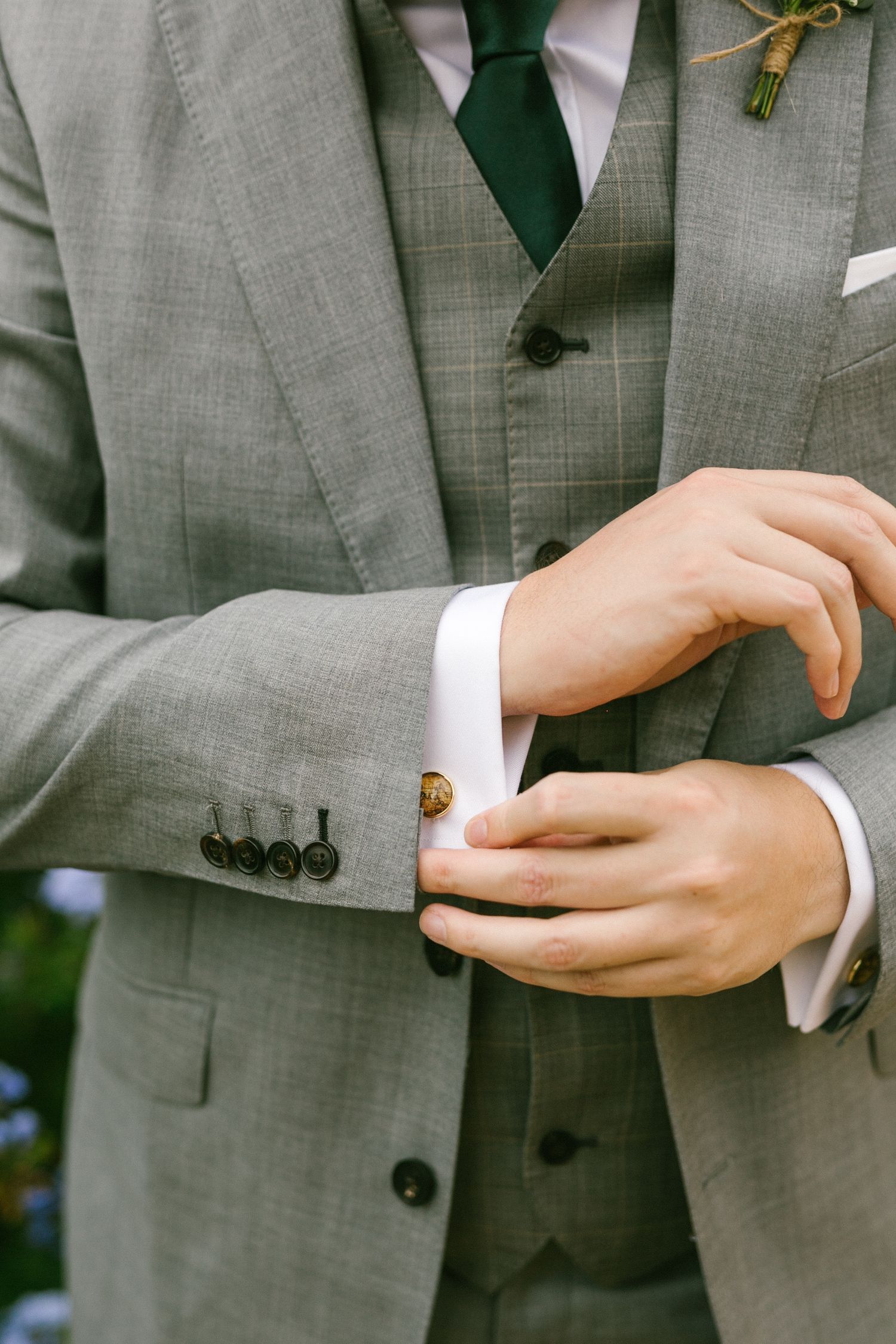 Close-up of a grooms hands adjusting sleeve cuff on grey suit with green tie and white shirt.