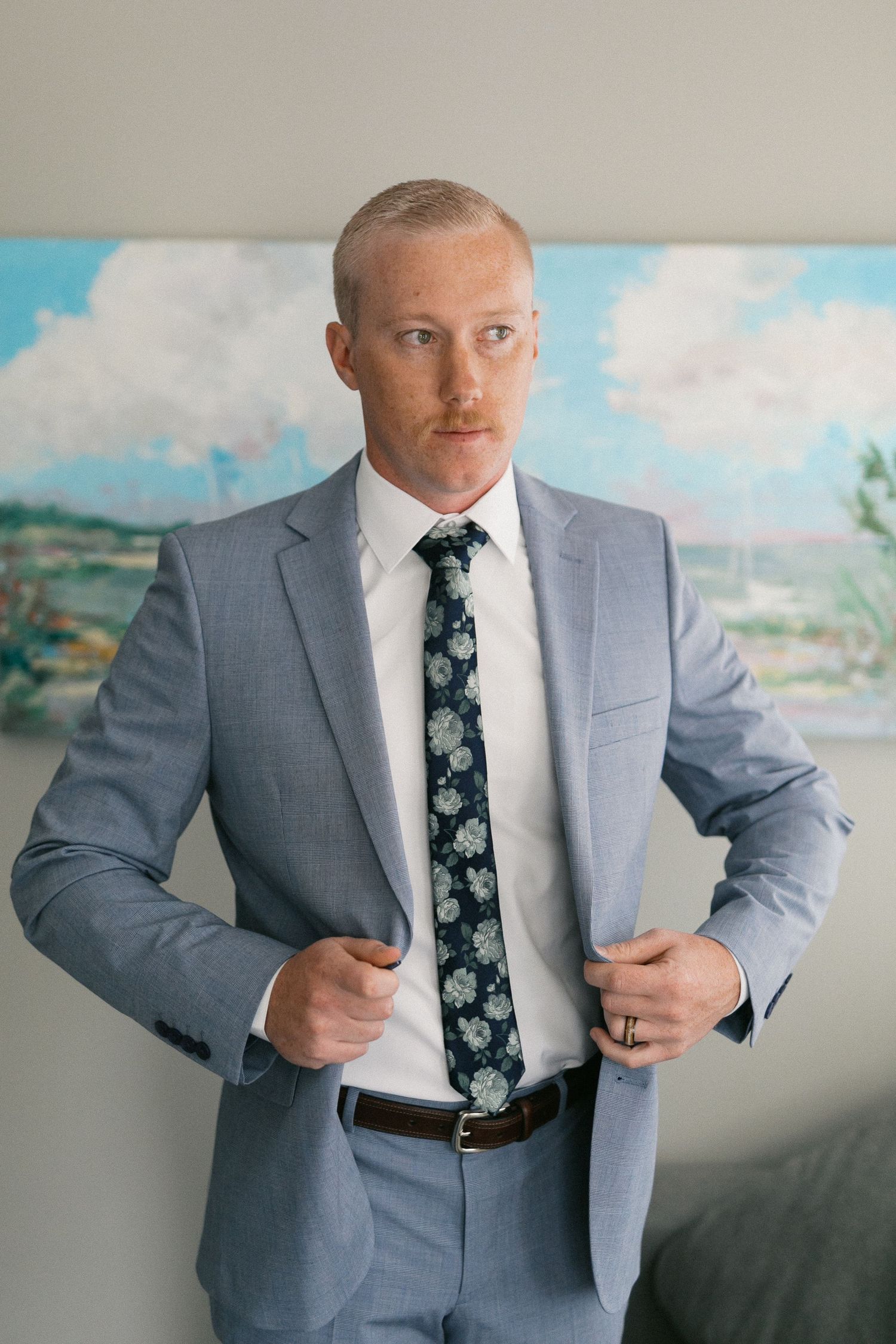 Groom adjusts his suit jacket and floral tie against scenic landscape backdrop.