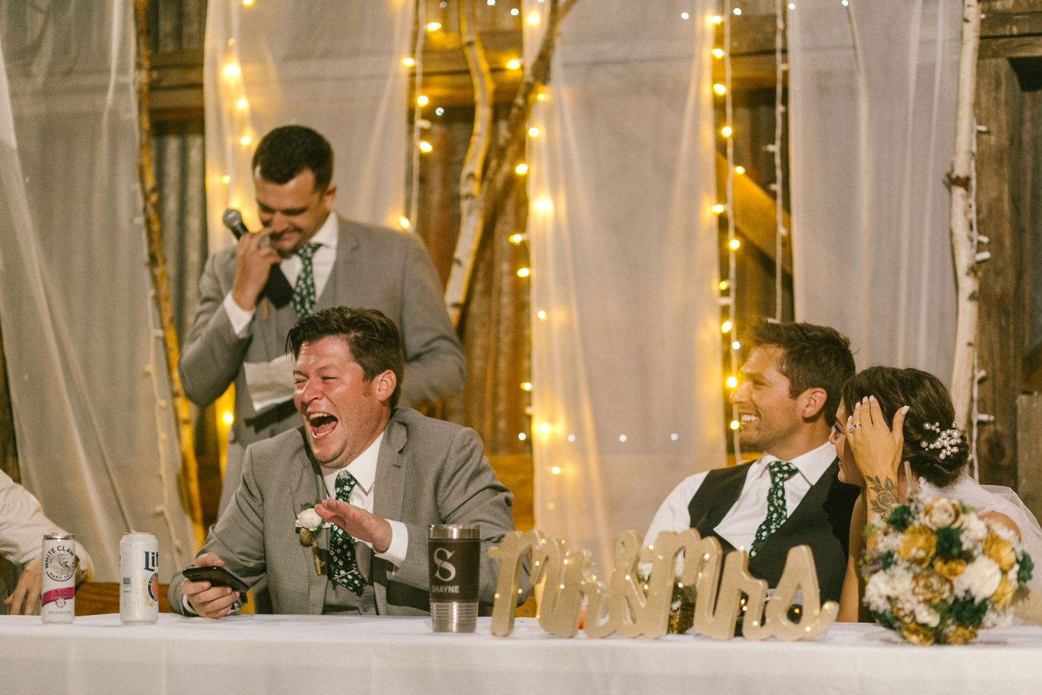Wedding reception guests laughing at head table decorated with golden lights and white draping.