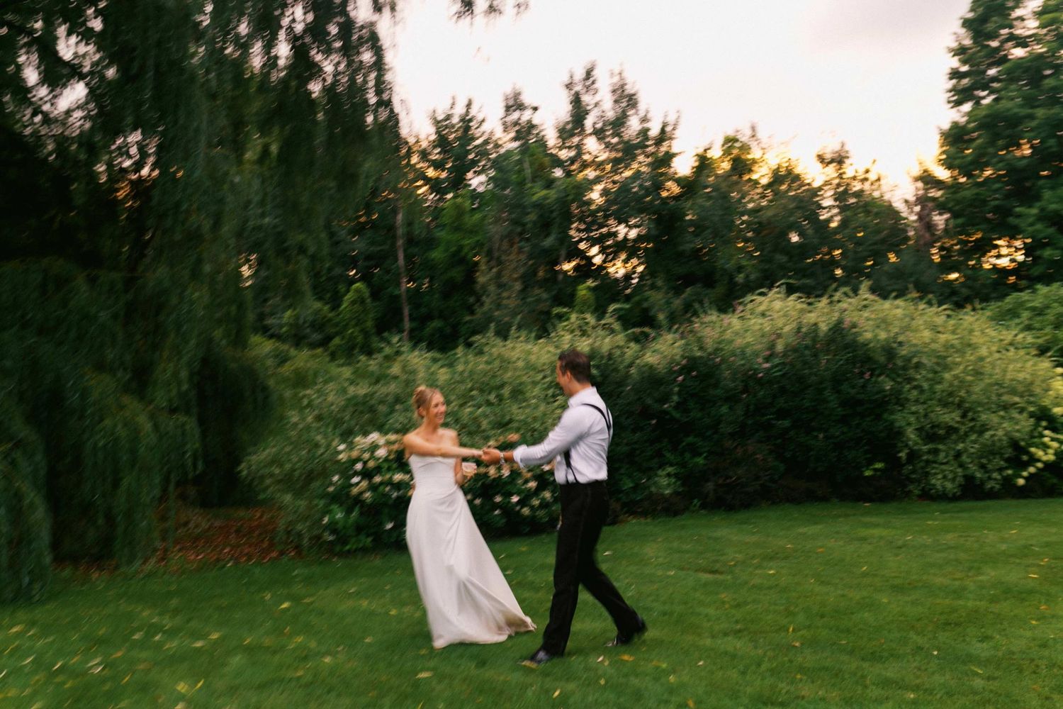 Couple in wedding attire walks through a lush garden setting during sunset golden hour.