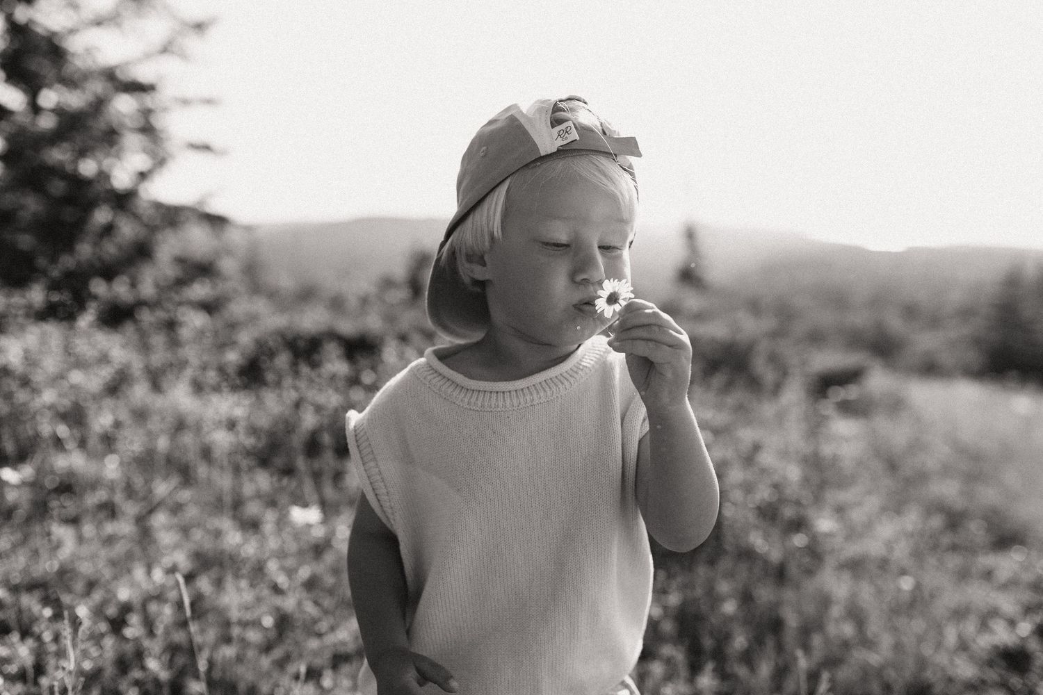 Black and white photo of a toddler in a cap smelling a small flower in an outdoor meadow.