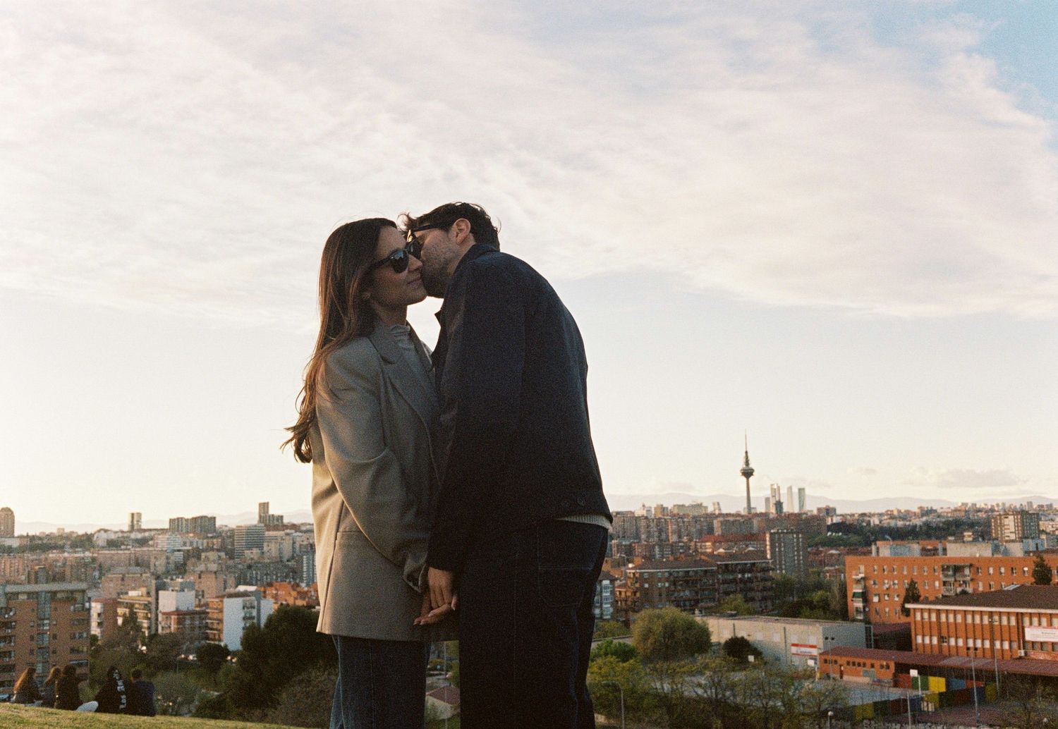 Couple kissing on hilltop at sunset with city skyline and TV tower visible in background.