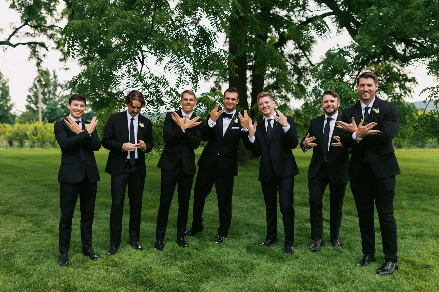 Groomsmen in black suits laugh together while holding drinks on a lush green lawn during outdoor wedding celebration.