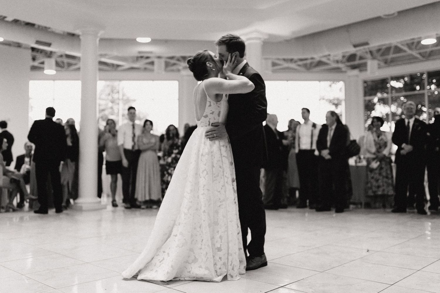 Wedding couple dancing together in an elegant white ballroom during their first dance as guests watch from the sides.