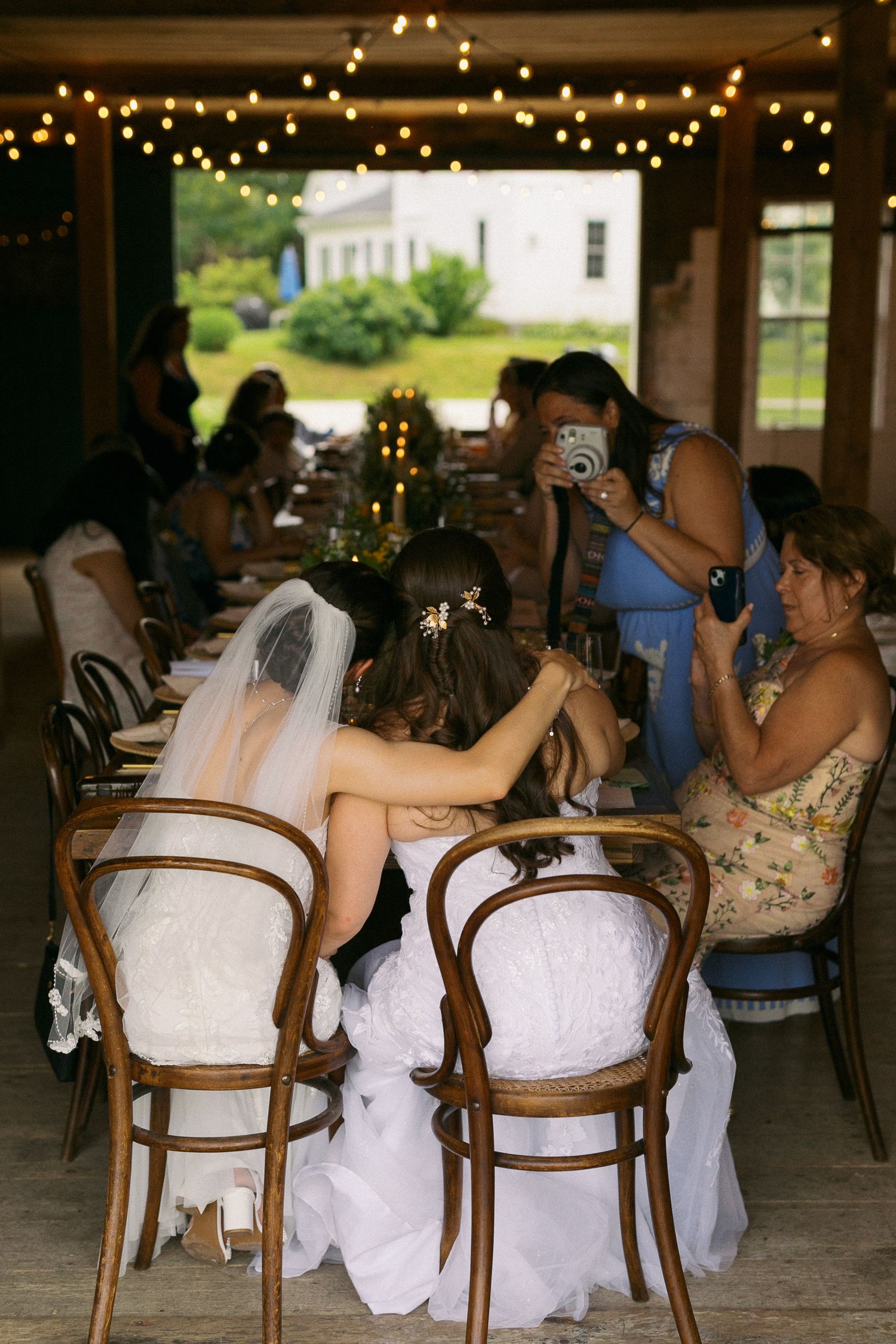 Bride in white gown and veil seated at wedding reception table with guests and string lights overhead.