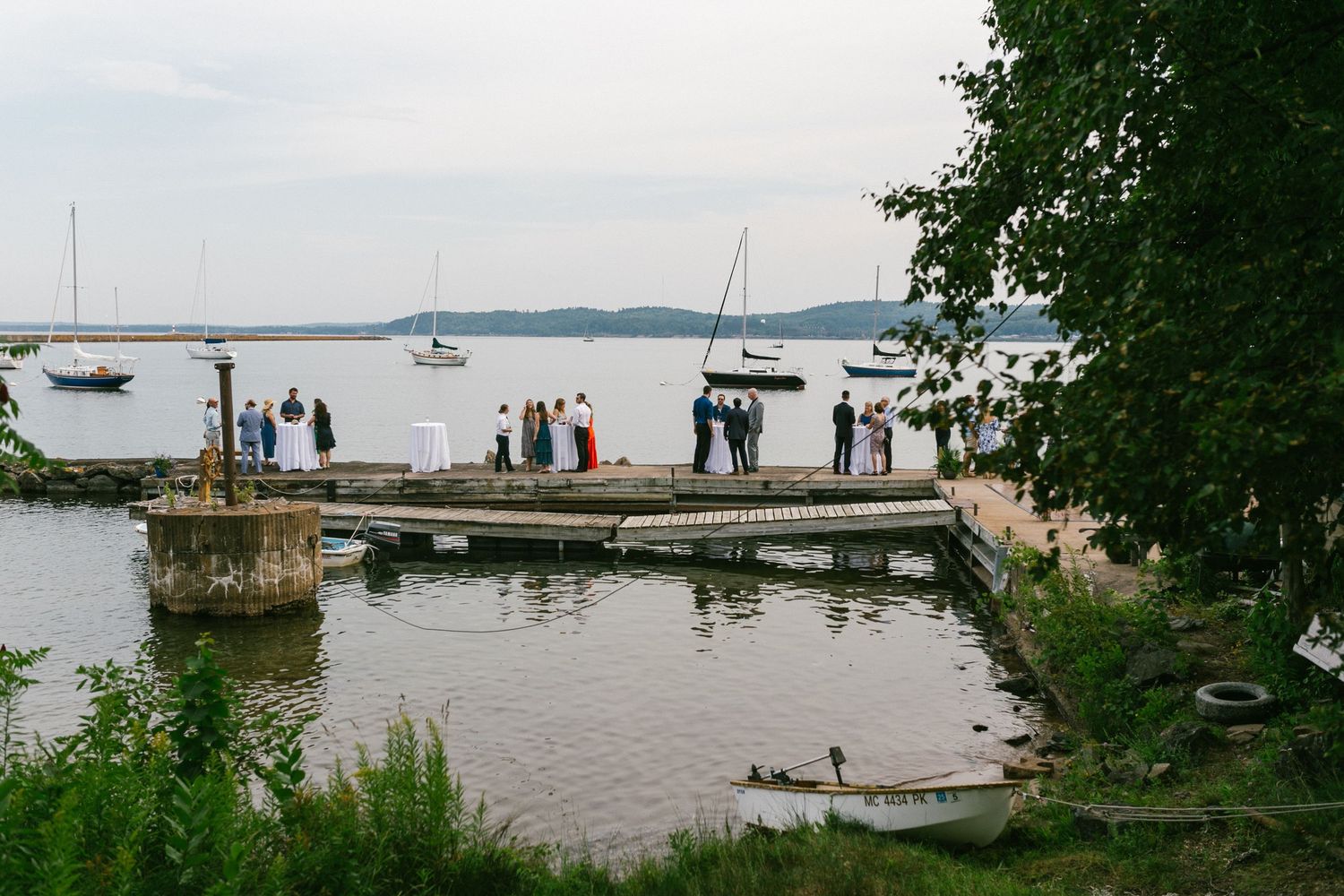 Scenic waterfront wedding venue with wooden dock, sailboats, and lush greenery along the shoreline.