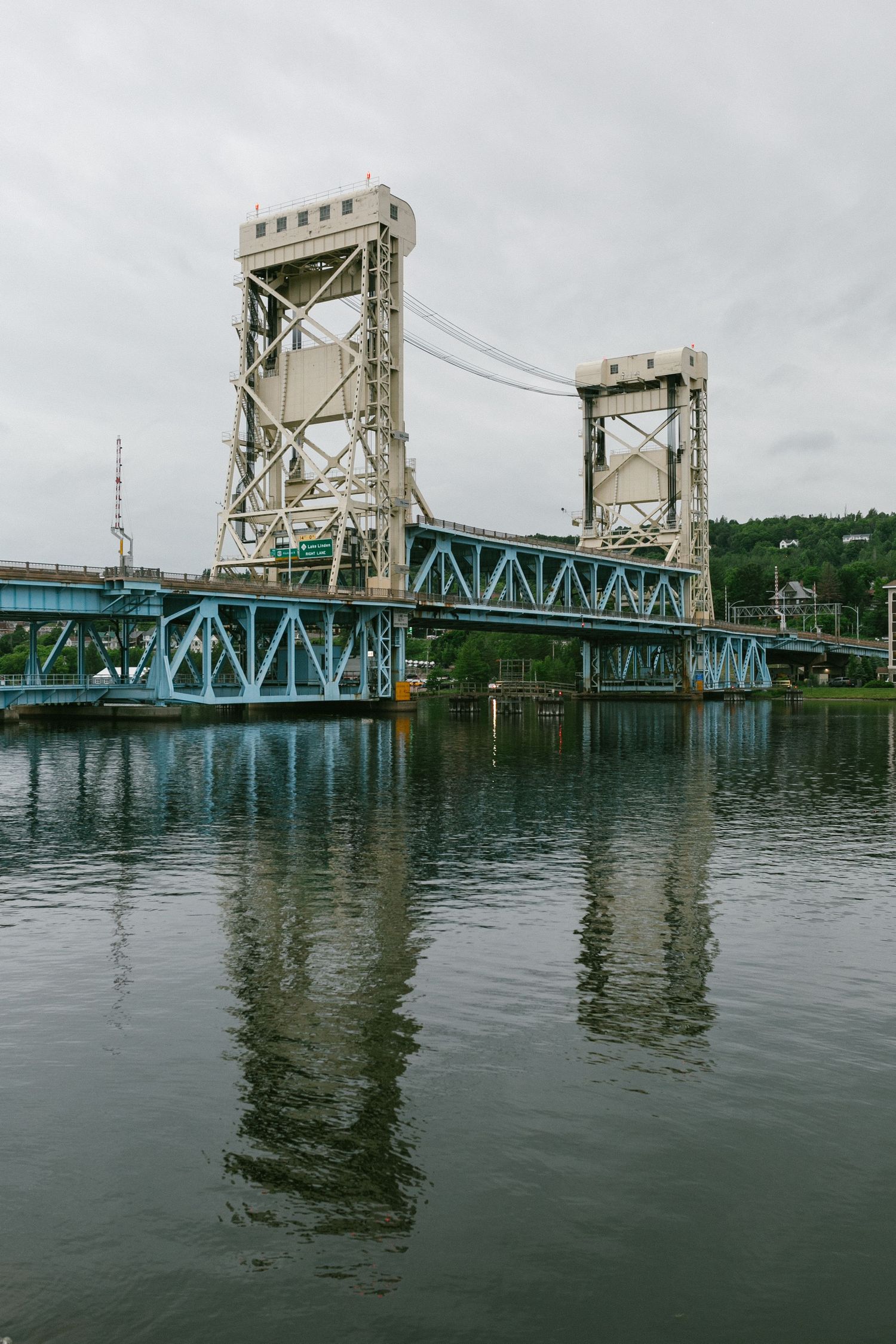 A vertical lift bridge with blue steel beams and concrete towers reflects in calm waters under cloudy skies.