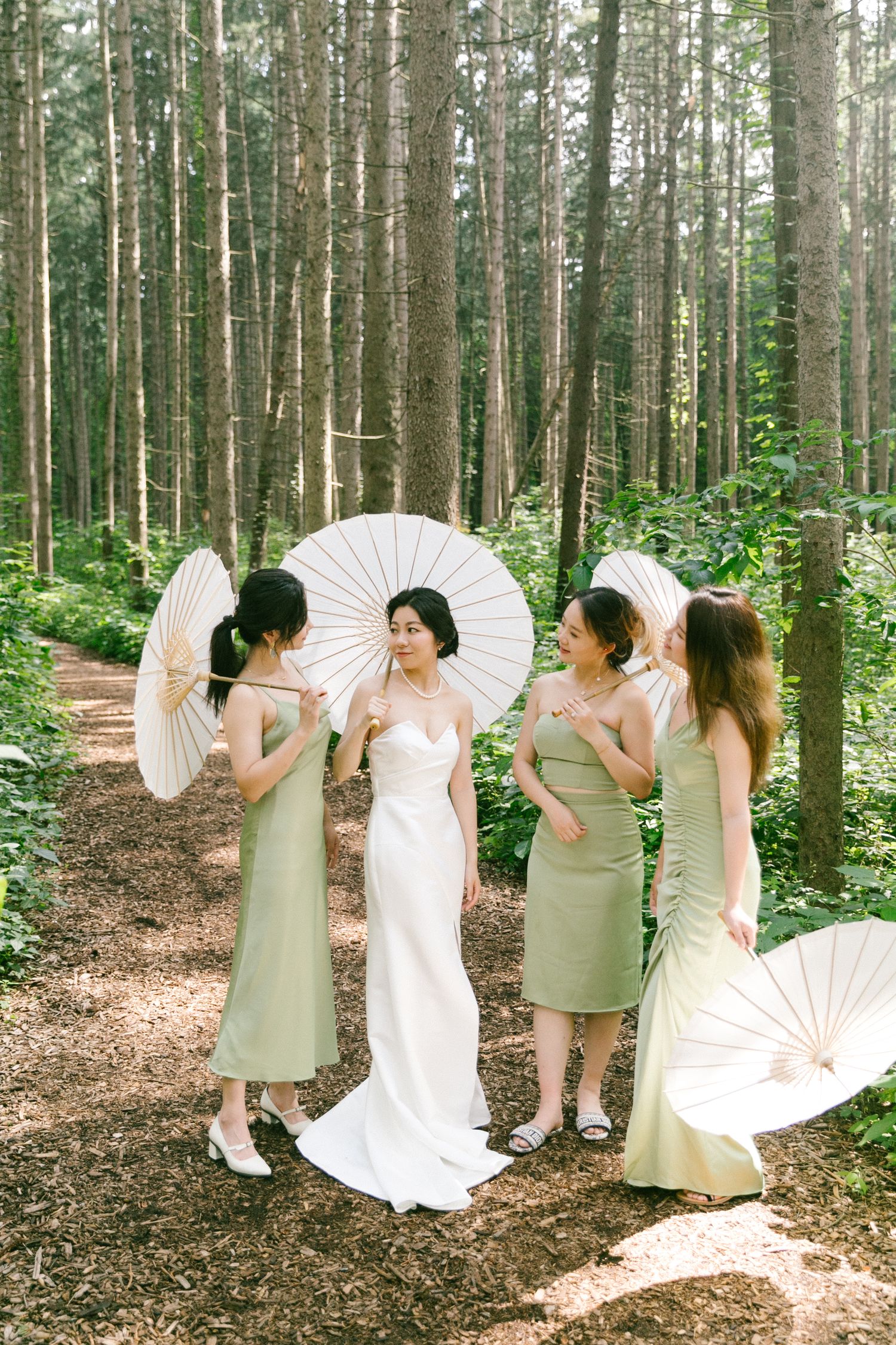 Bride in white gown poses with bridesmaids in sage green dresses holding white parasols in forest.