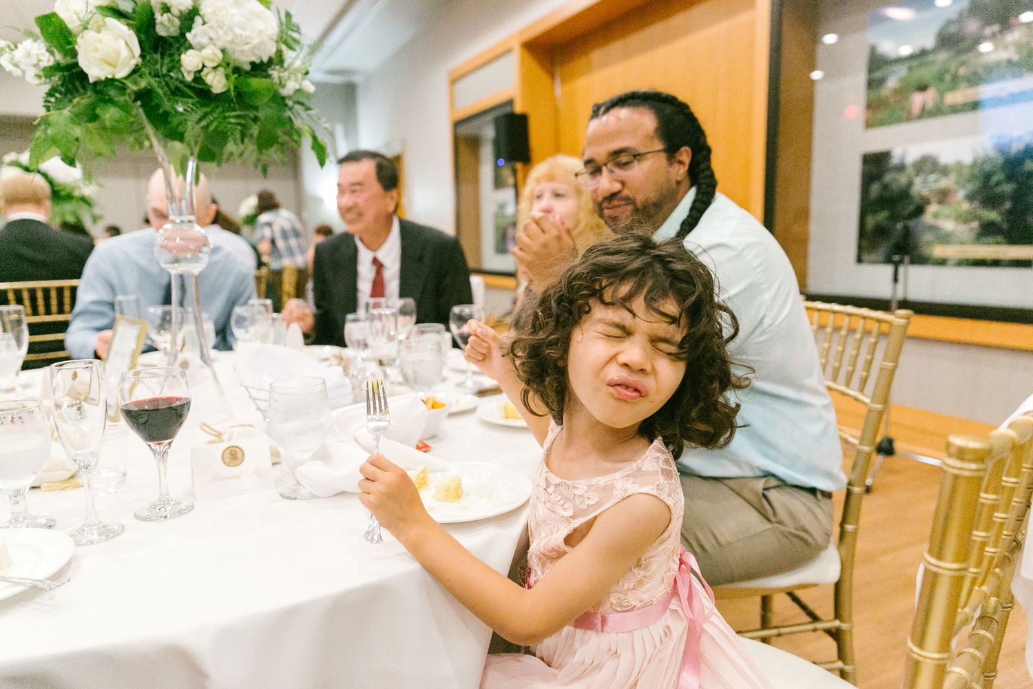 Young girl in pink dress sits at elegant wedding reception table with man behind her, floral centerpieces visible.