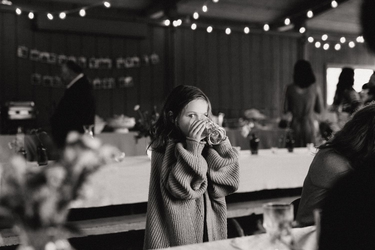 A black and white photo of a young girl drinking from a large glass at a reception with string lights in the background.