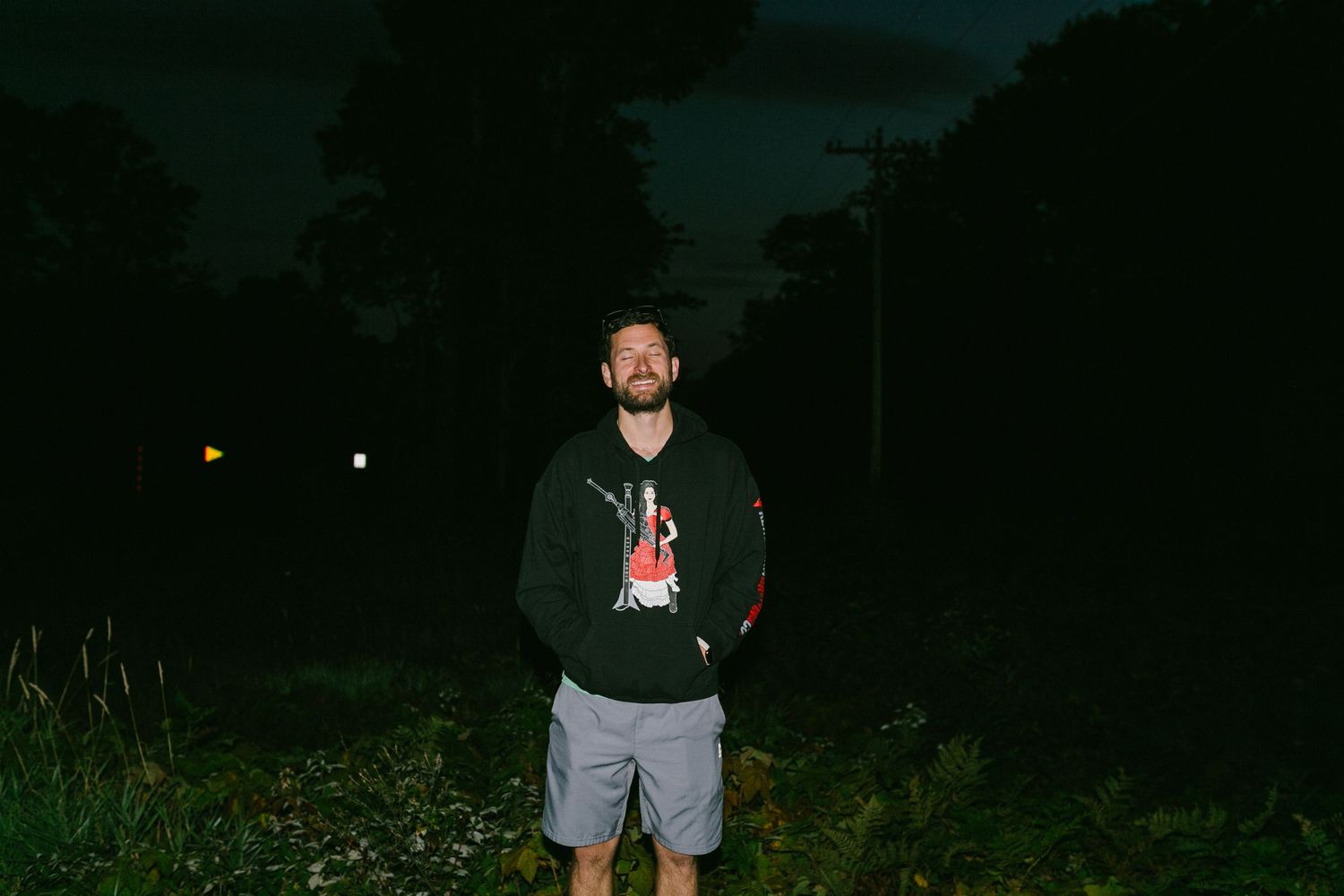 A groom wearing a black hoodie and gray shorts stands outdoors at night in a dark nature setting.
