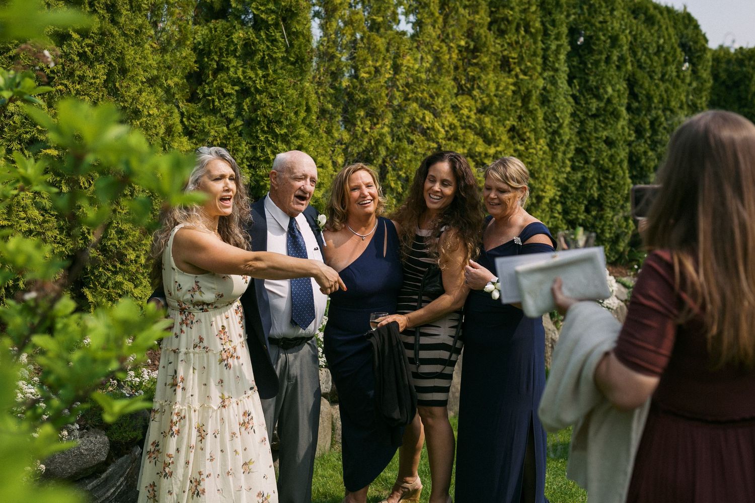 Wedding guests mingling outdoors among lush green trees and gardens.