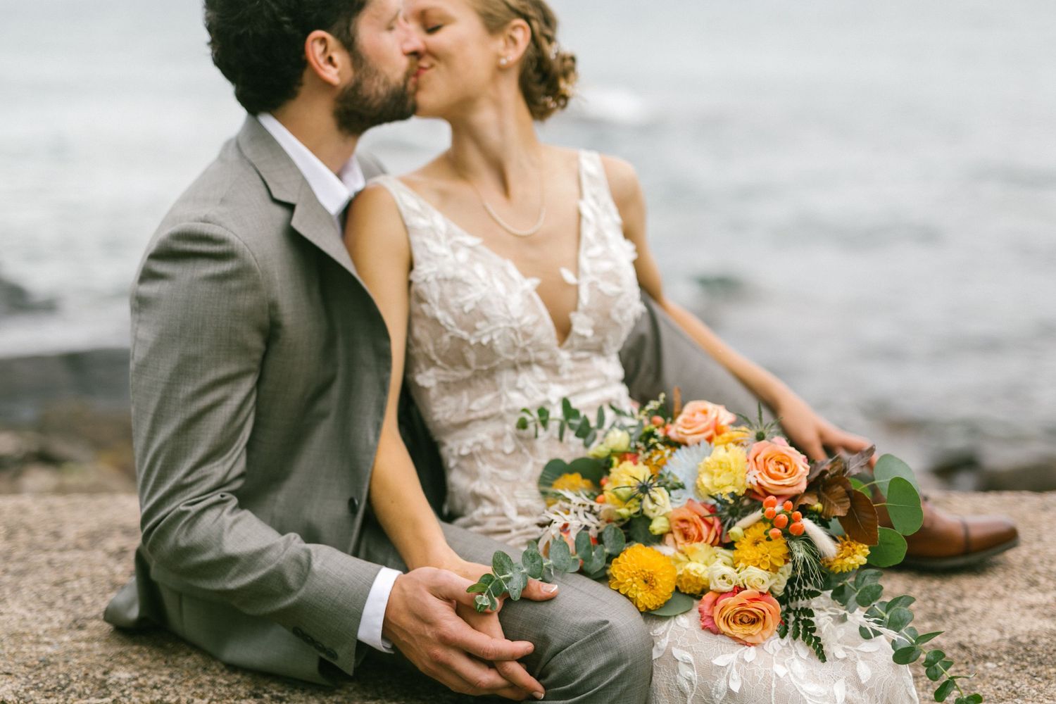 Couple in wedding attire share romantic moment on rocky shore with vibrant yellow and orange bouquet.