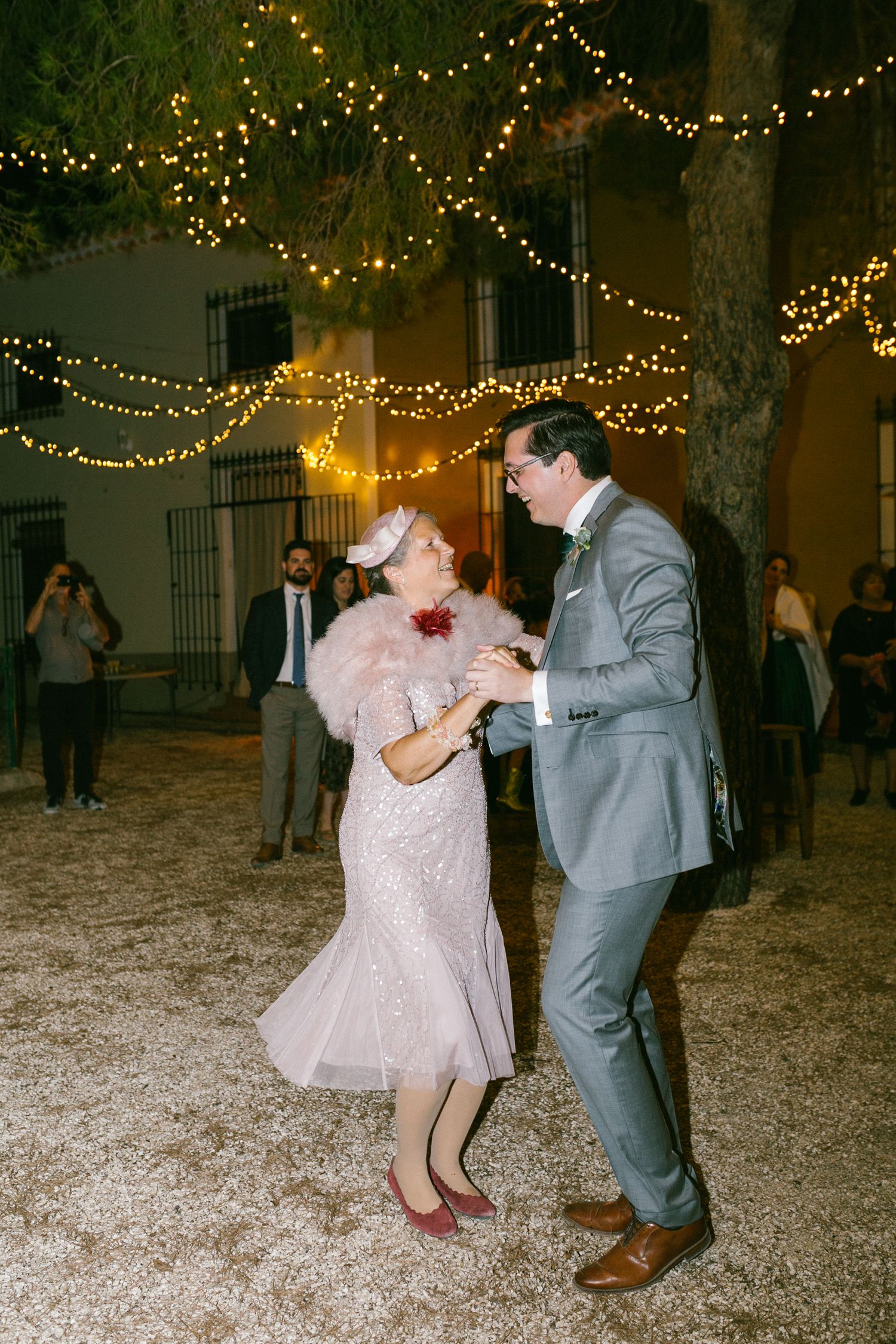 A couple dances together under twinkling string lights at an outdoor evening wedding reception.