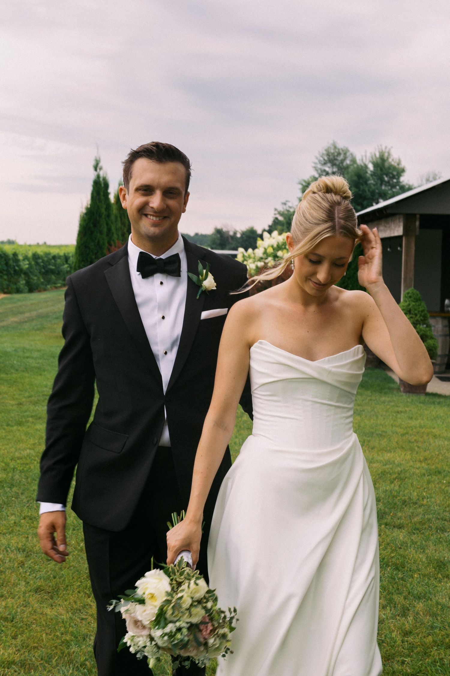 A couple in wedding attire walks on a green lawn, with the bride in white holding a bouquet and groom in black tuxedo.