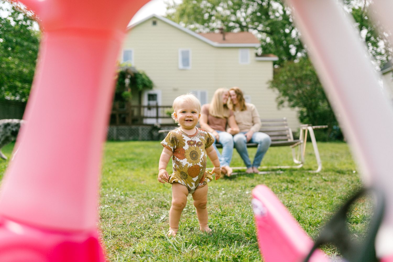 Smiling toddler in floral outfit playing in backyard while parents relax on bench.