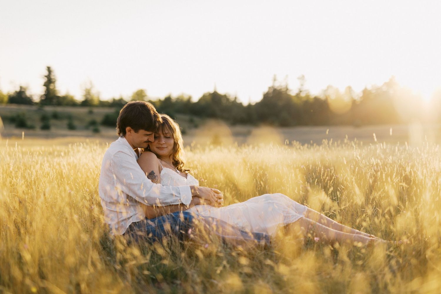 Couple sitting together in golden field at sunset, sharing a romantic embrace in tall grass.
