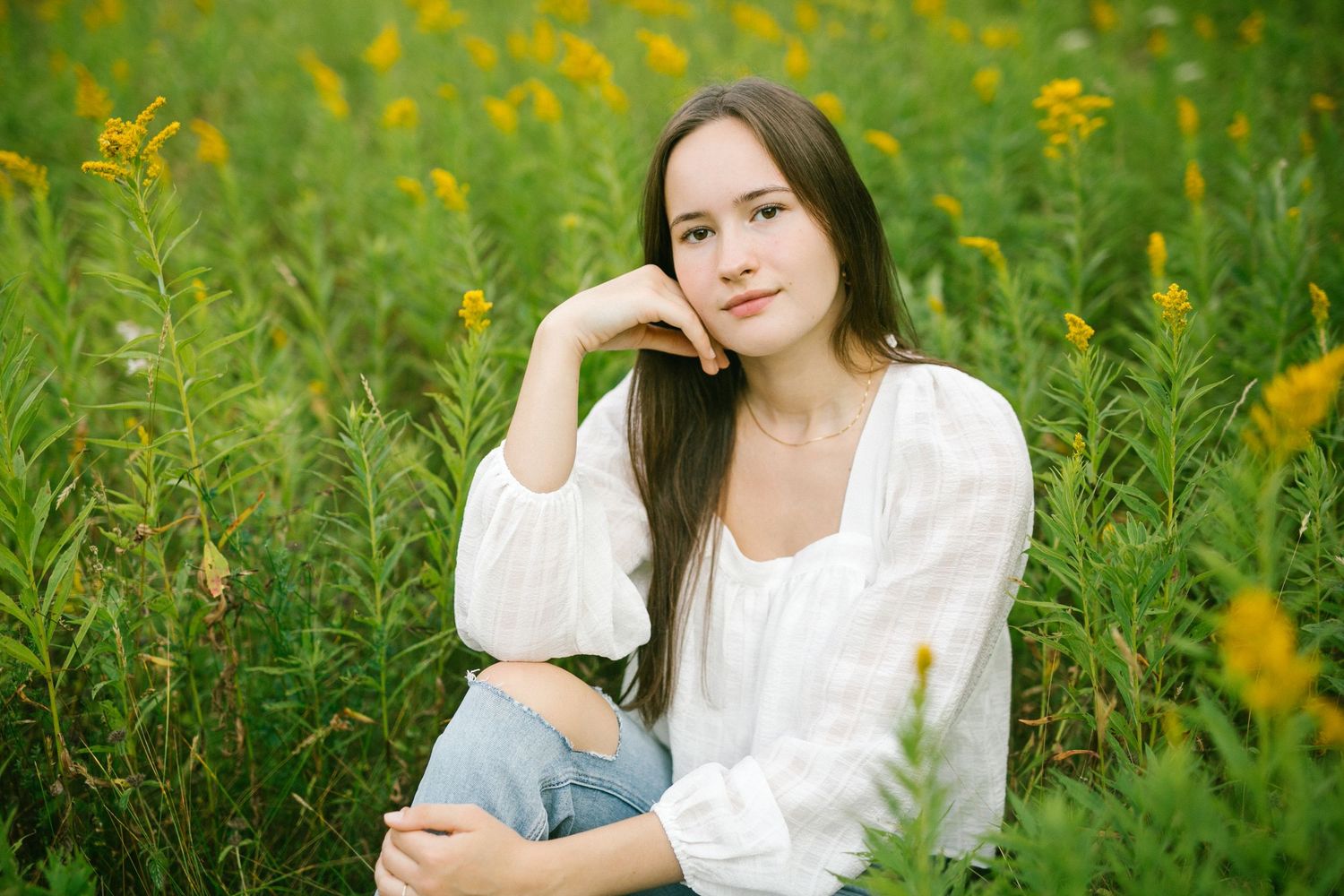 Young woman in white blouse sitting among yellow wildflowers in a lush green field.