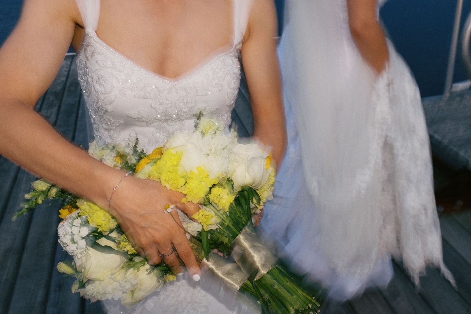 Bride in white lace gown holding yellow and white wedding bouquet with ribbon wrap on steps.
