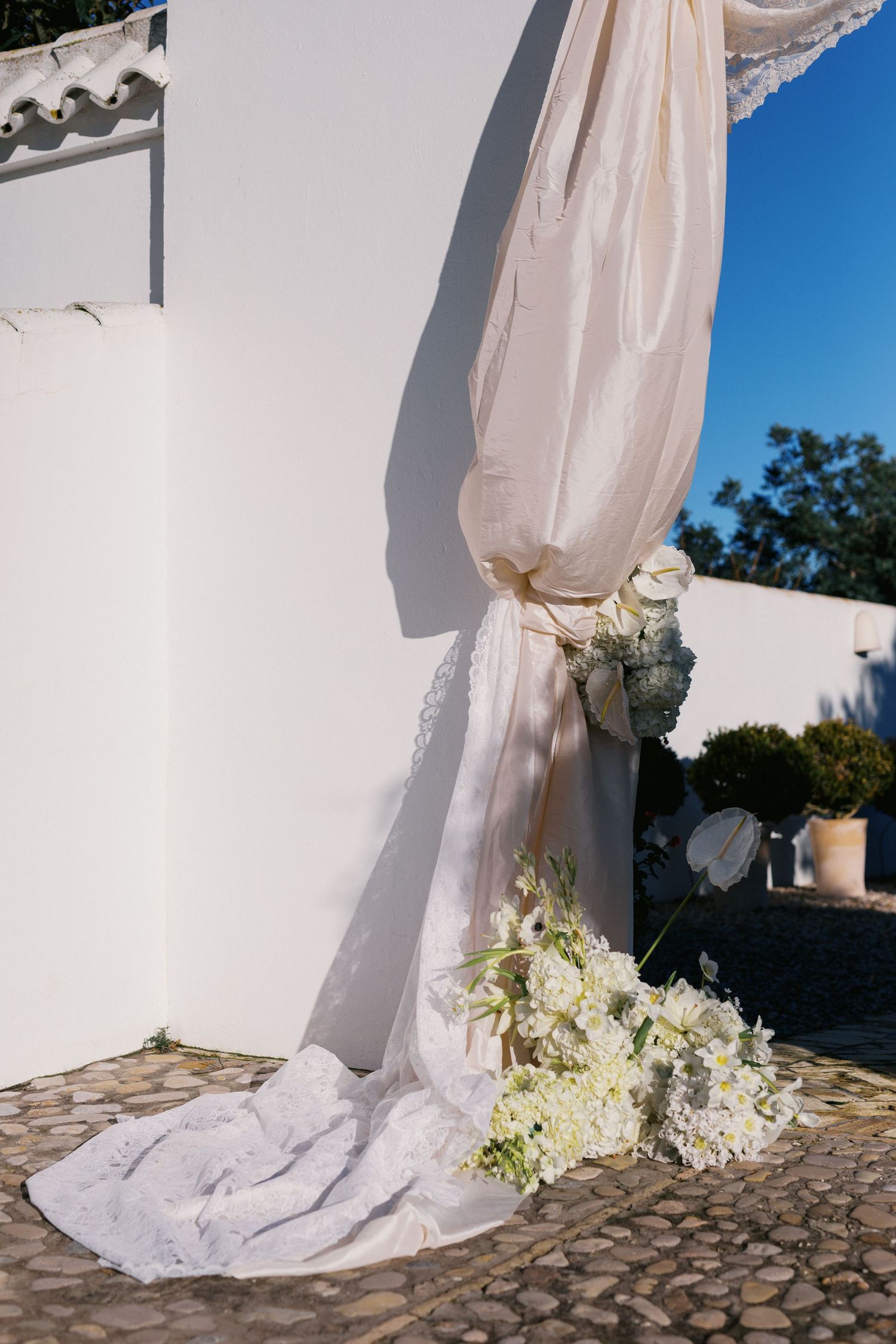 Elegant wedding arch decoration with cream fabric draping and white hydrangea bouquet against white wall.