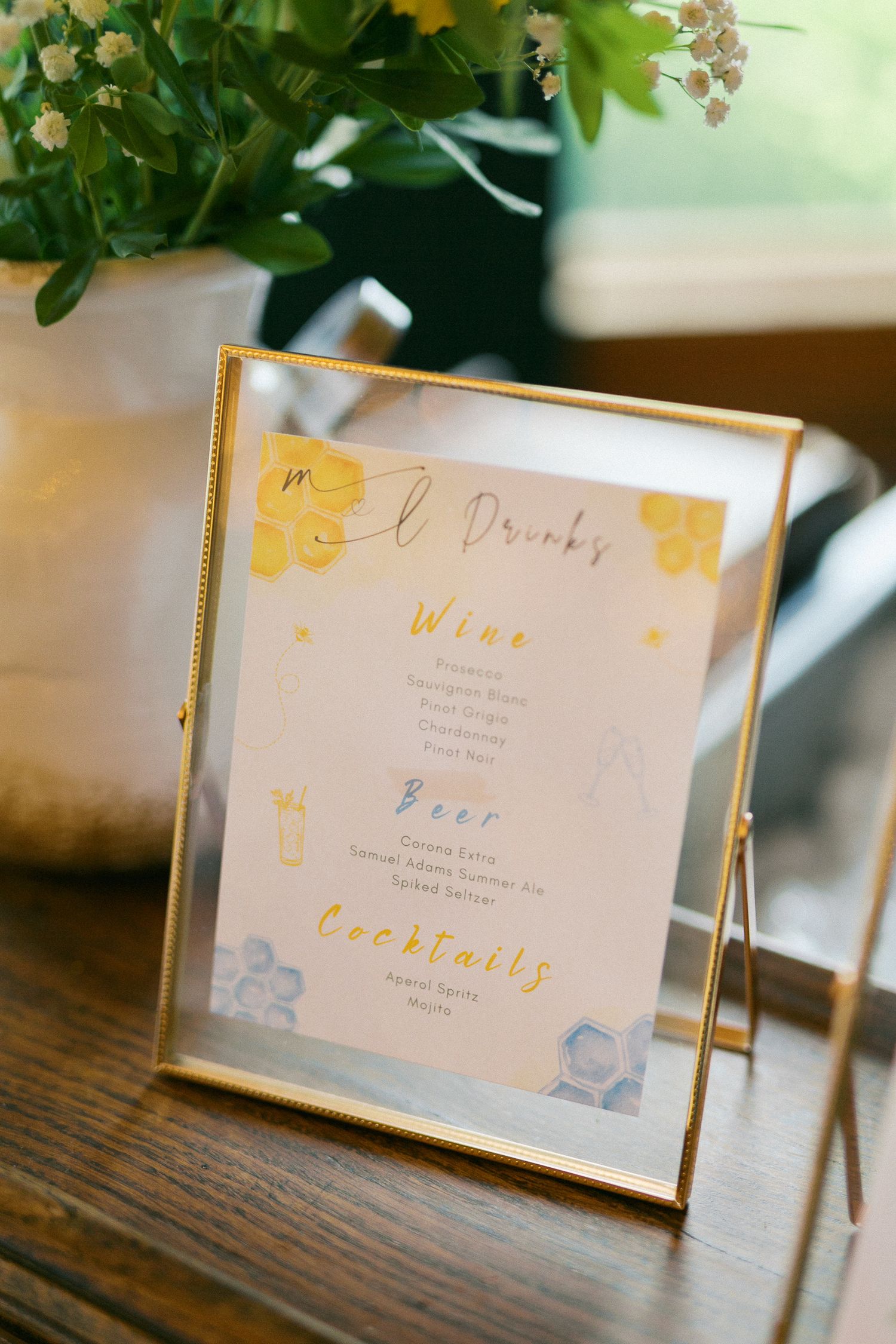 Gold-framed wedding bar menu displaying wine, beer, and cocktails with yellow floral accents on a wooden table.
