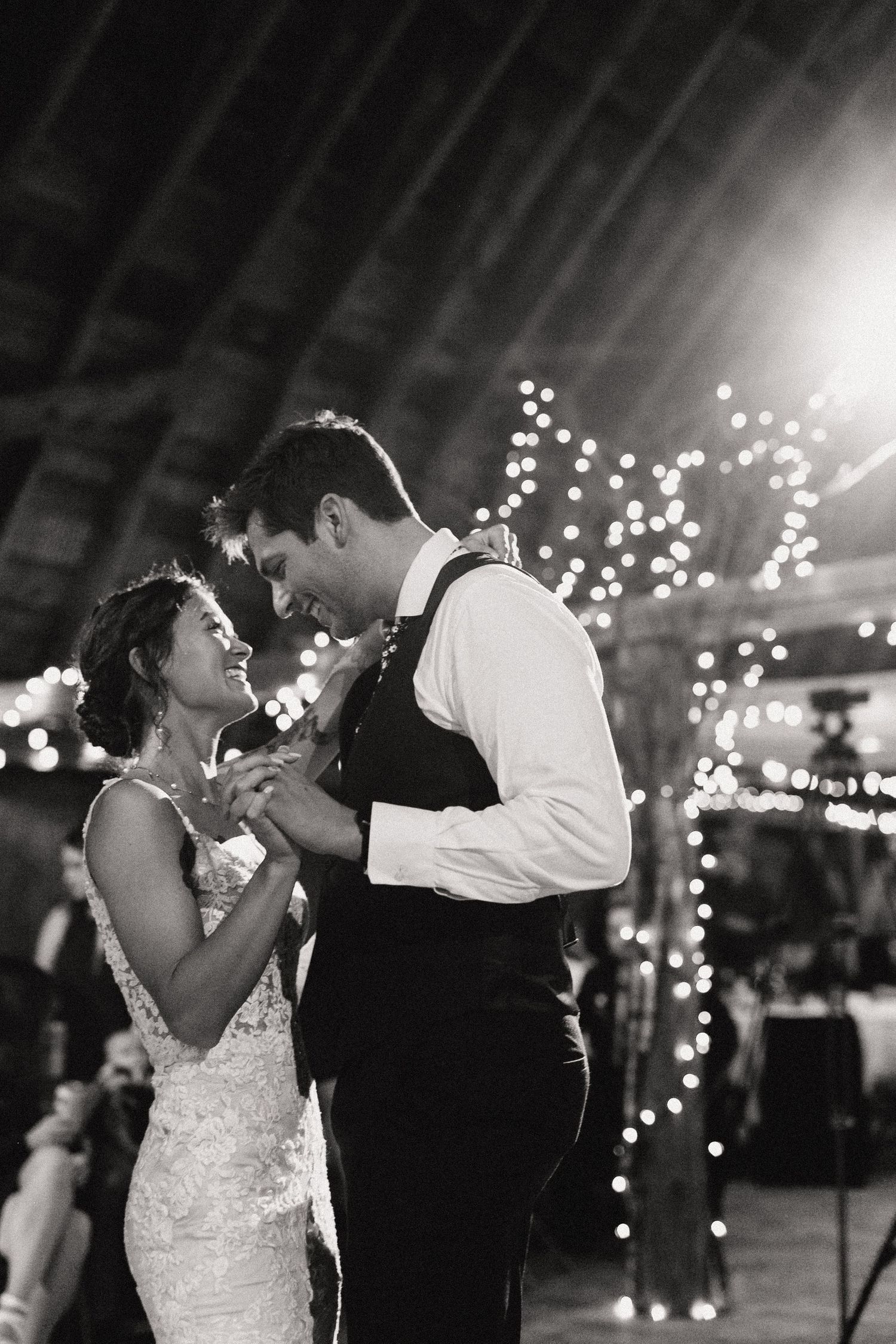 A couple shares an intimate first dance moment at their wedding reception under twinkling string lights in black and white.