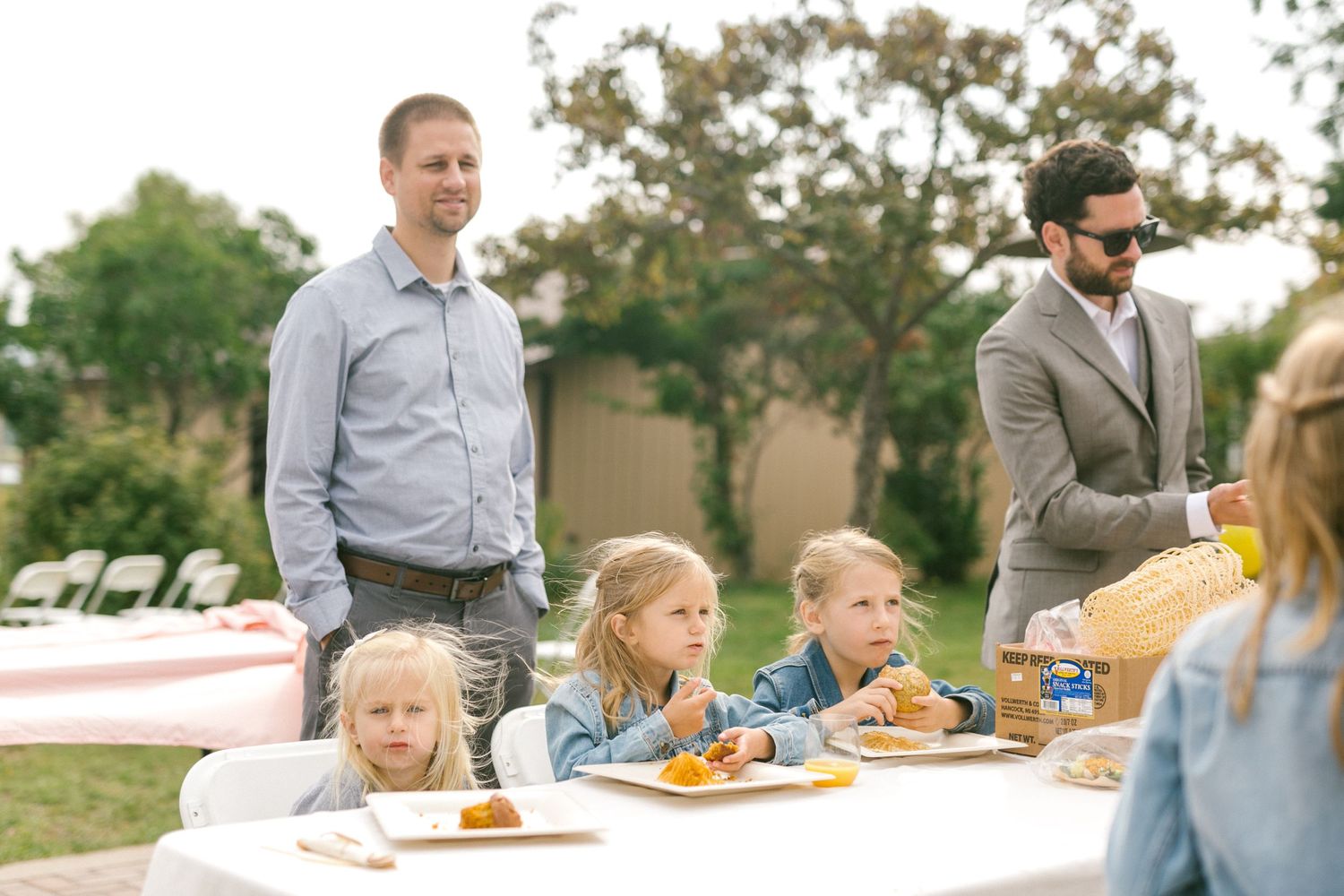 People gather at outdoor table with food while children enjoy their meal in a sunny backyard setting.