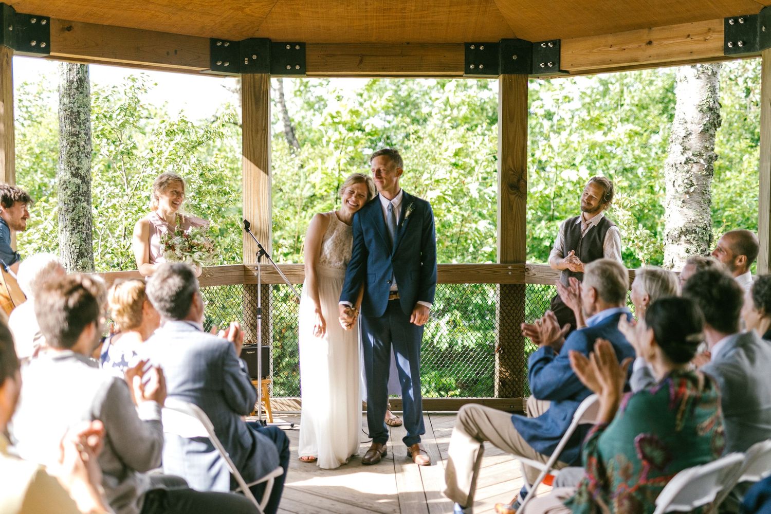 Sunlit outdoor wedding ceremony in wooden gazebo with seated guests and natural forest backdrop.