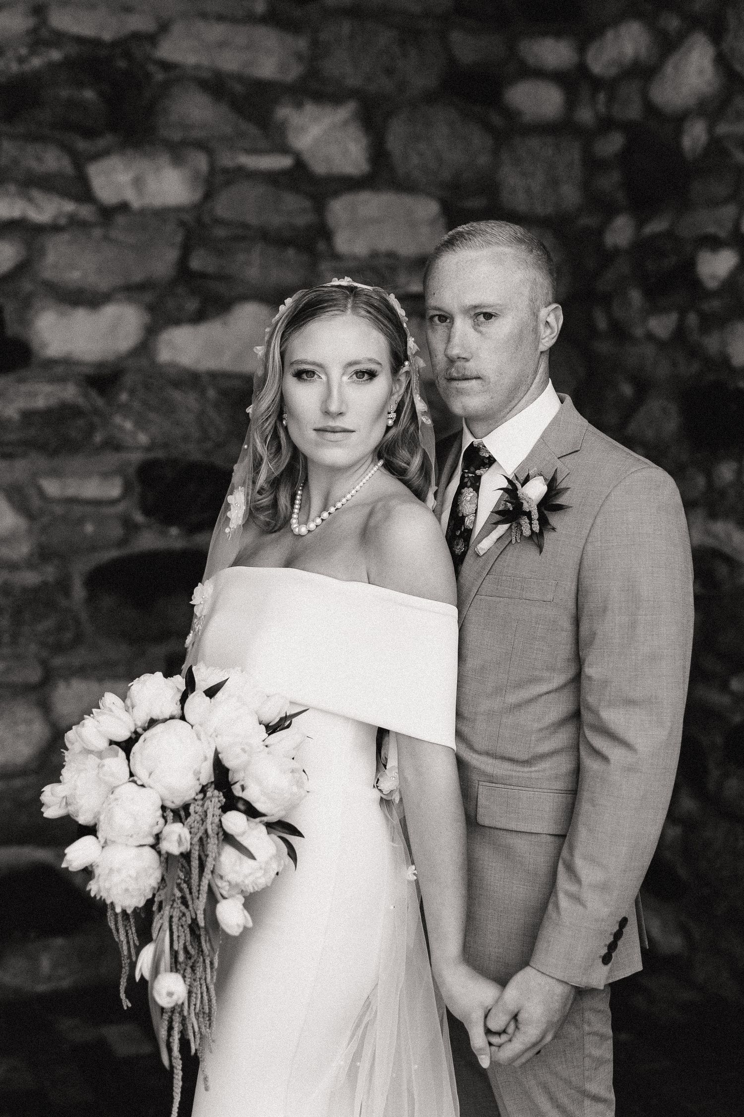 Black and white wedding portrait against rustic stone wall with white floral bouquet.