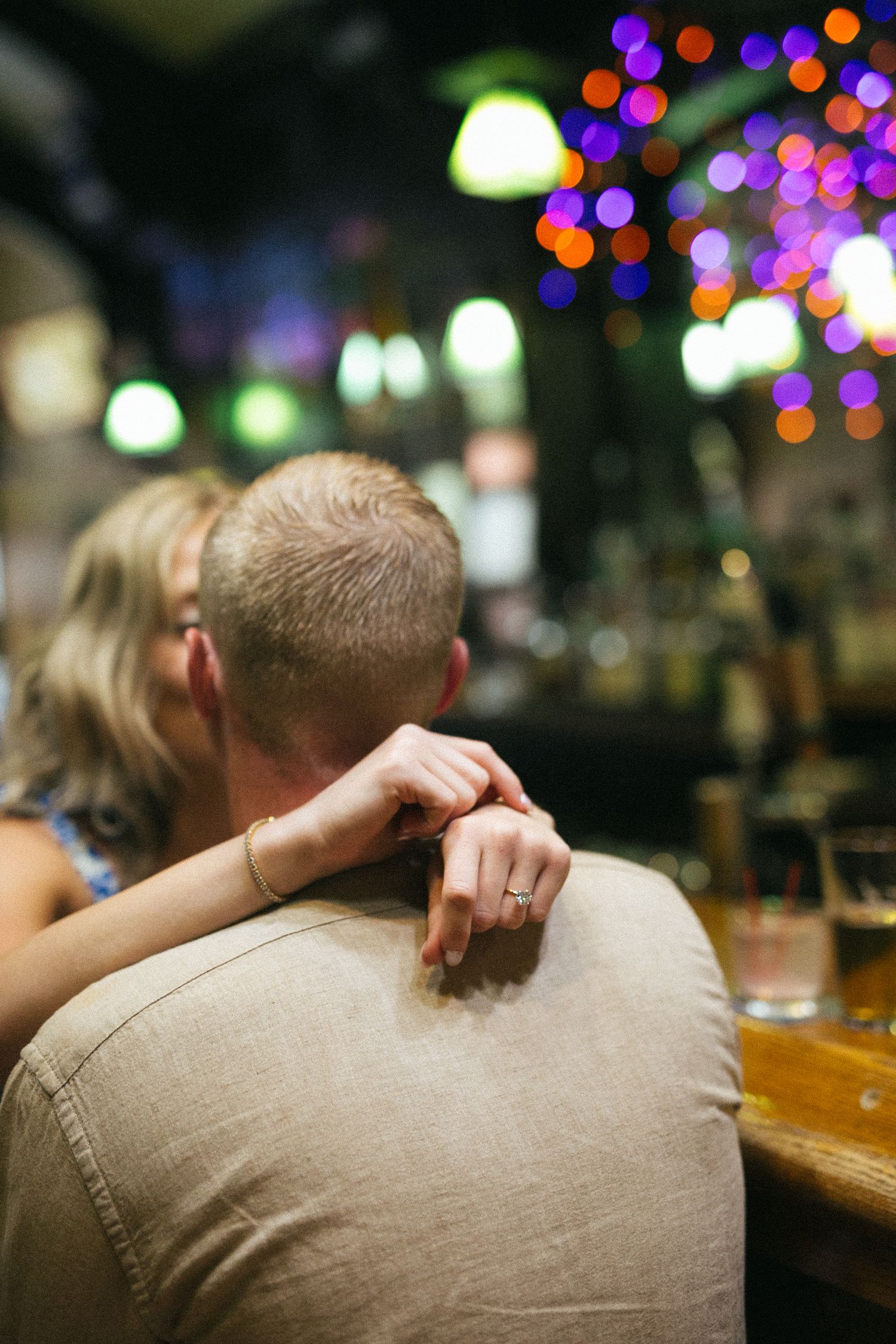 A couple sits at a bar with colorful bokeh lights in the background while sharing an intimate moment.