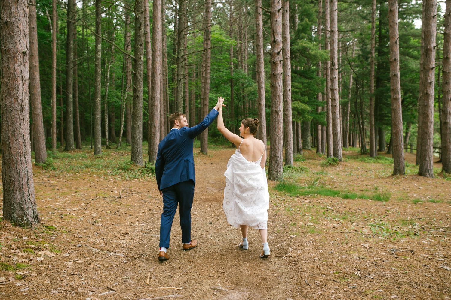 Two people in formal attire hold hands while walking down a forest path surrounded by tall pine trees.