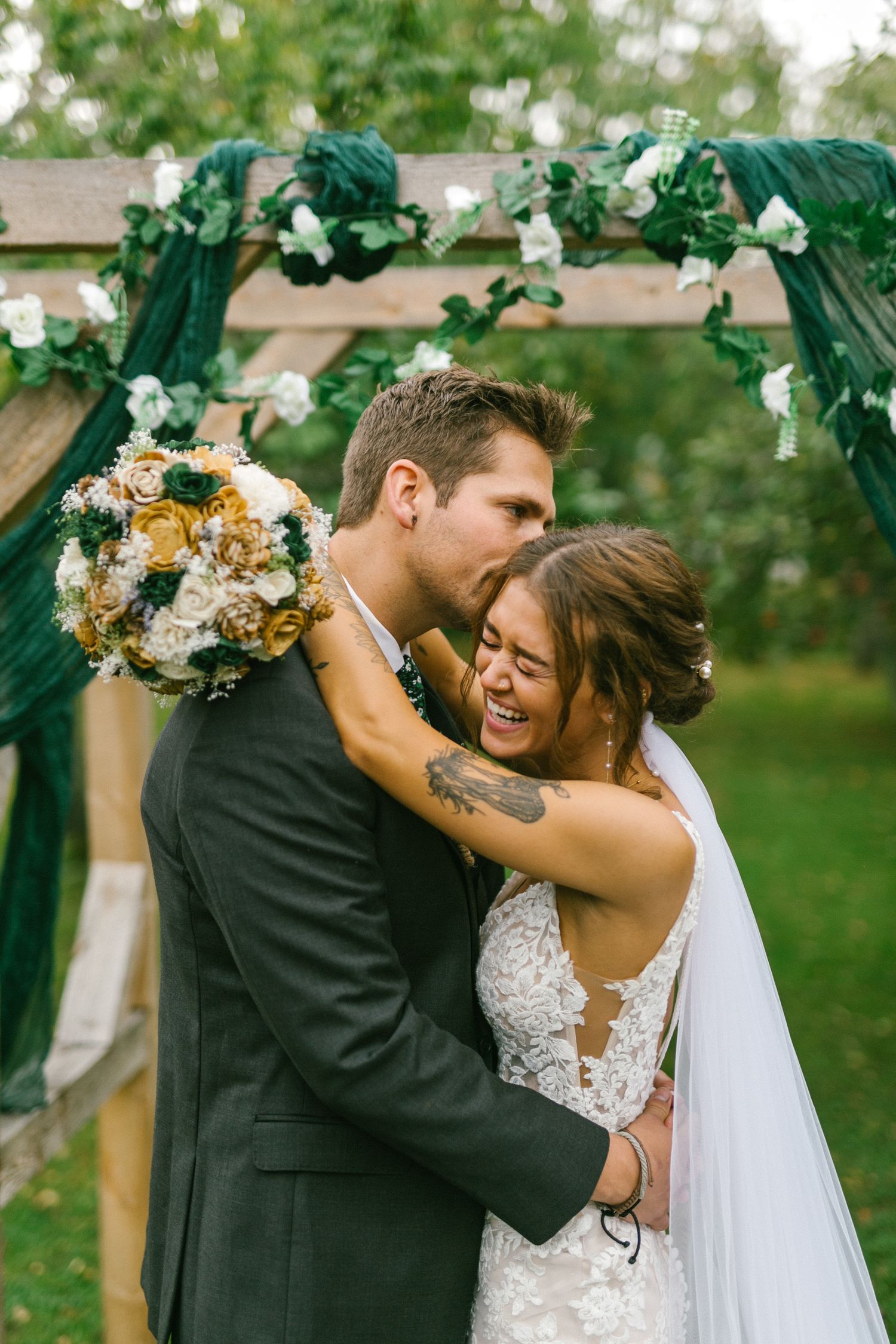 Newlyweds share a joyful moment under a greenery-decorated arch at an outdoor wedding ceremony.