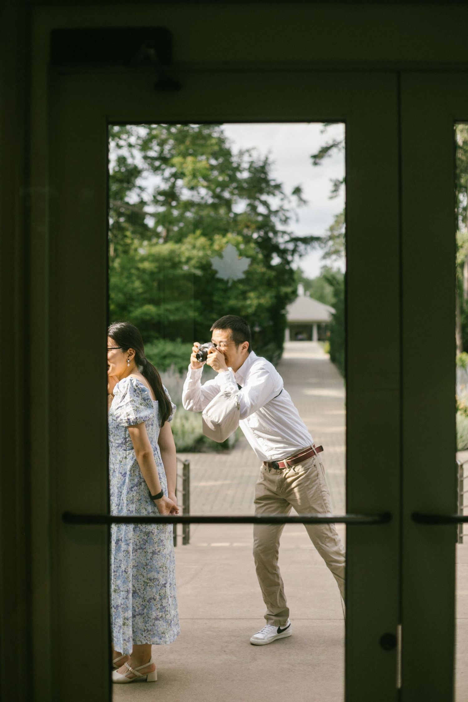 Man photographing with camera outdoors while woman in floral dress walks nearby, viewed through glass doors.