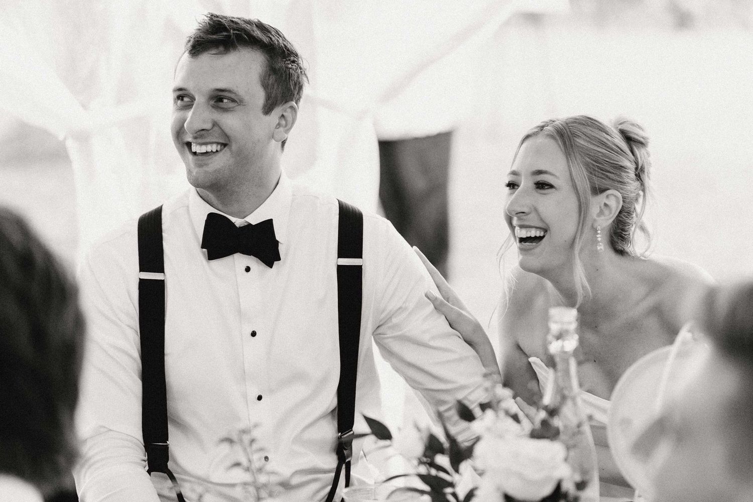 Black and white candid photo of a couple laughing together at their wedding reception, with the groom wearing suspenders.