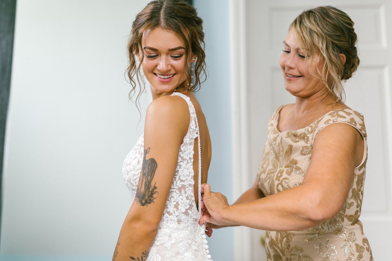 A bride and her mother share a joyful moment while getting ready in formal attire, with one helping adjust the other's white lace dress.