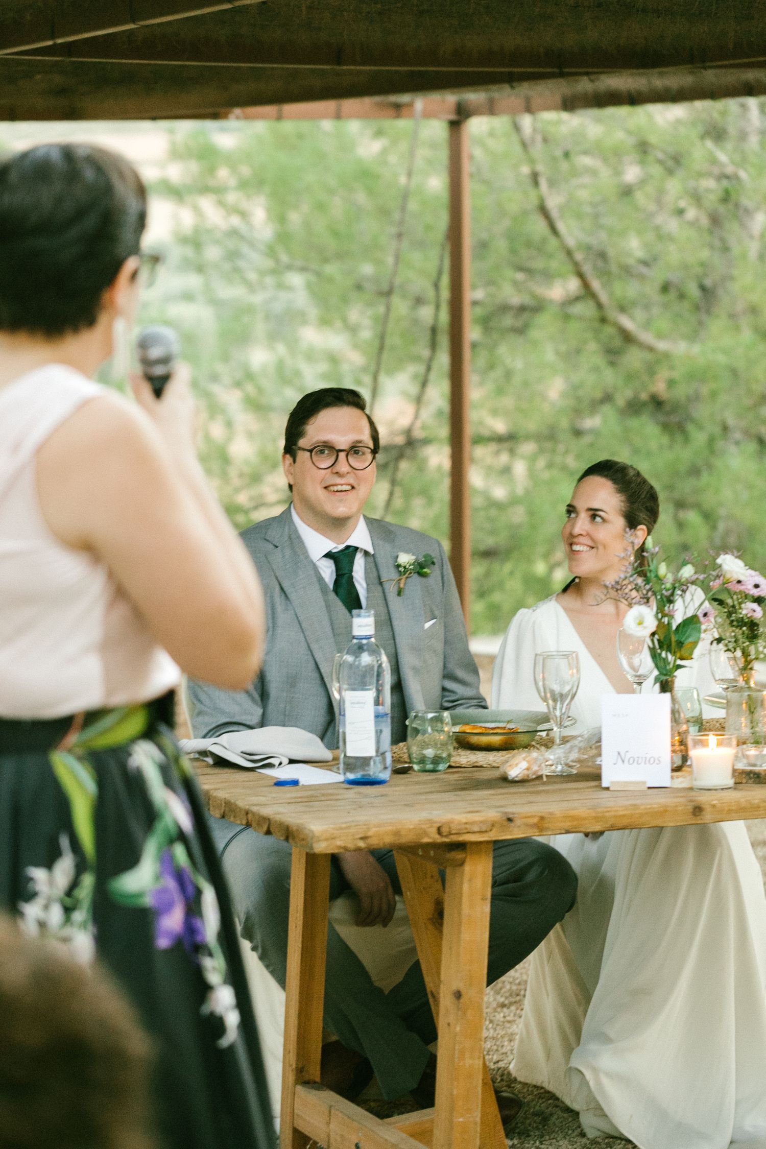 Wedding reception guests seated at rustic wooden tables under an outdoor canopy with floral centerpieces.