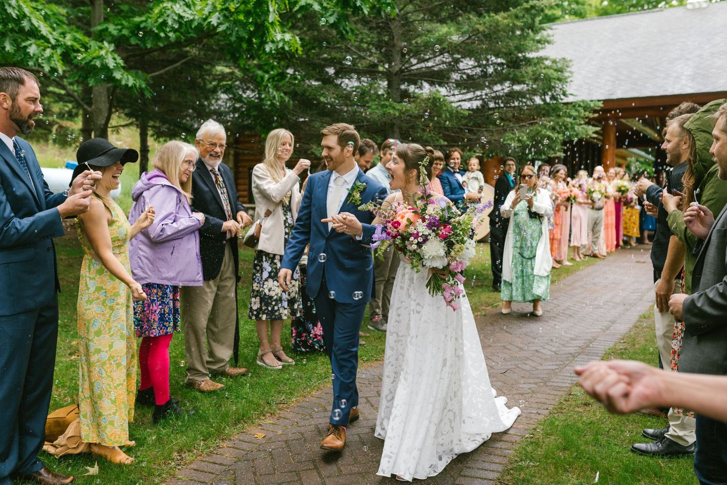 Wedding guests toss confetti as newlyweds walk down outdoor garden path surrounded by trees and guests.