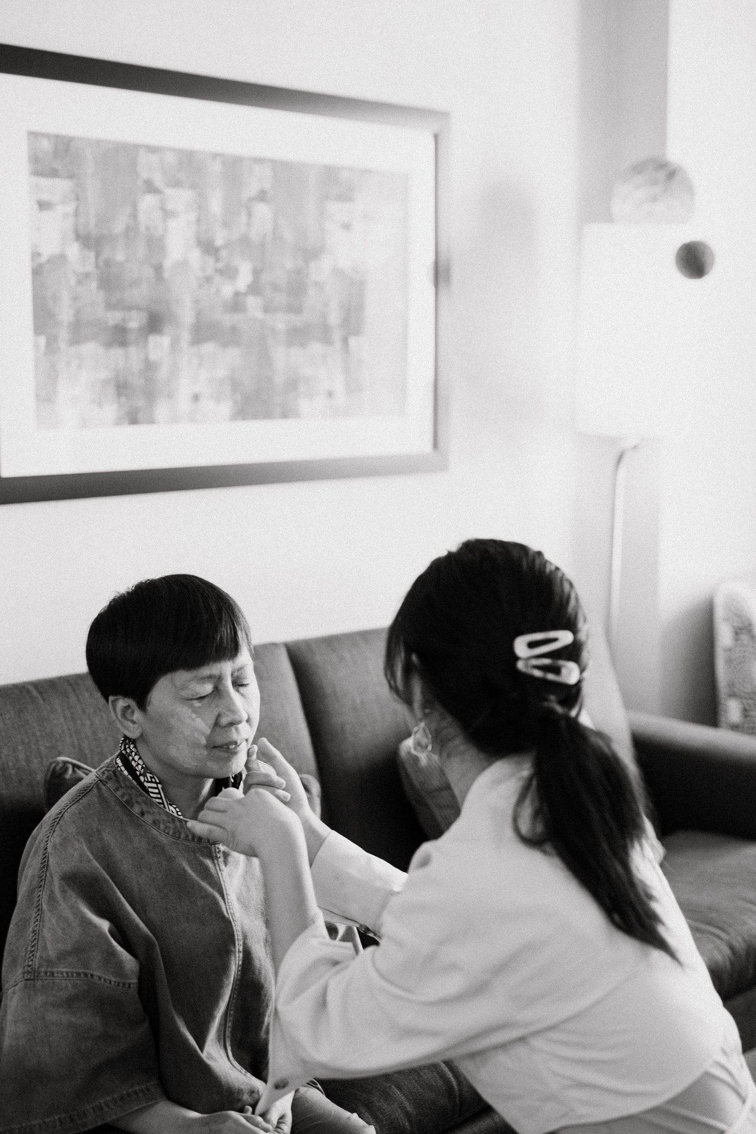 Black and white photo of a caregiver assisting an elderly woman on a sofa, with framed art on the wall behind them.