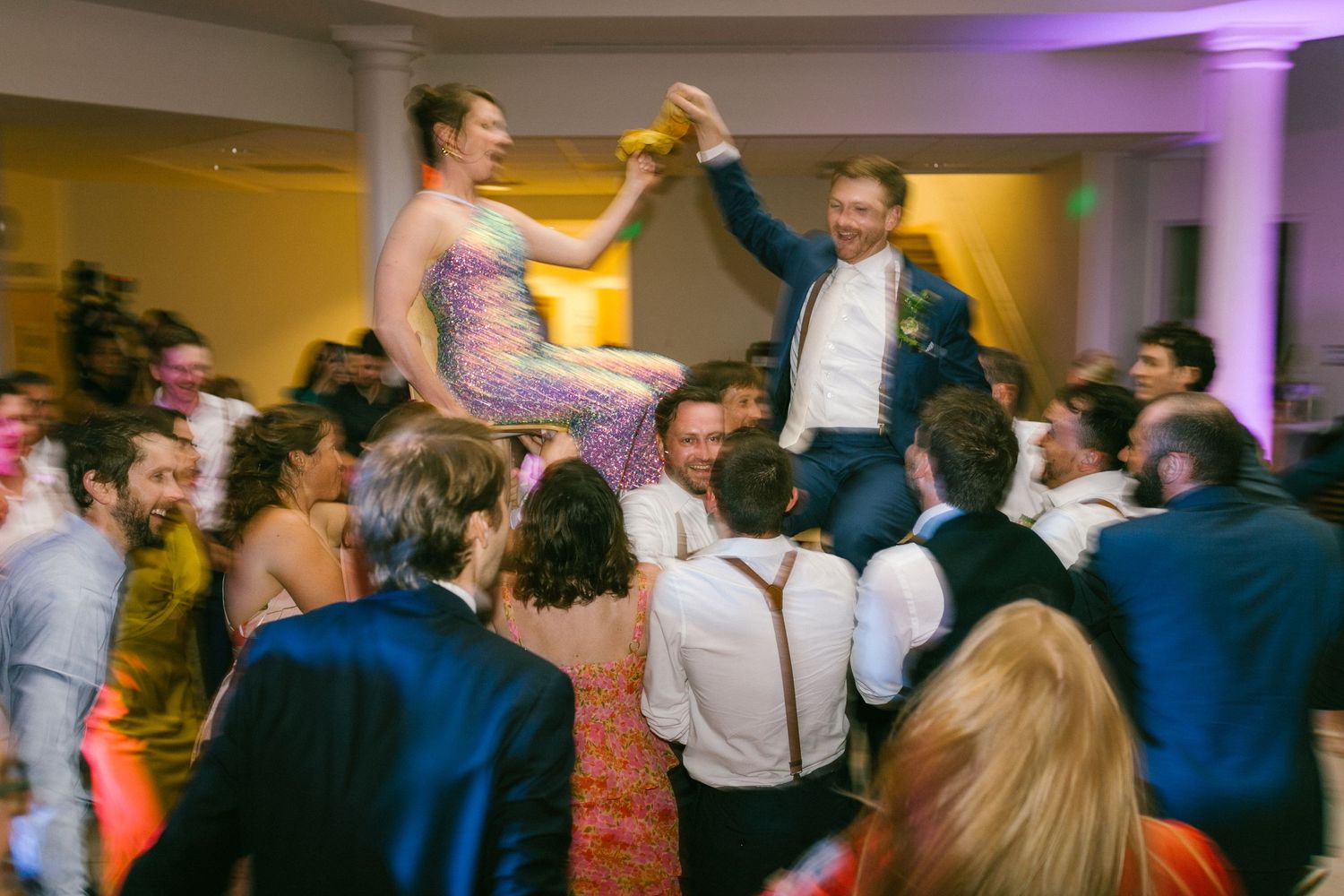 Wedding party guests dance and celebrate on a dance floor with purple mood lighting at a reception.
