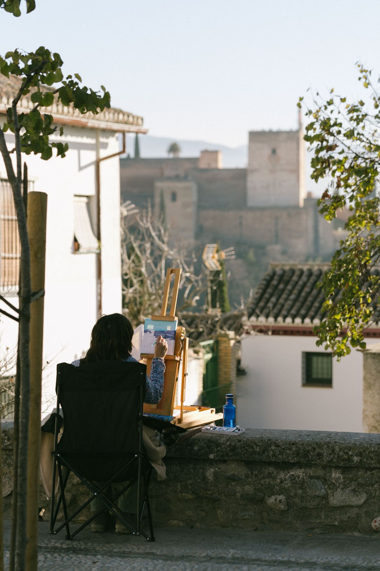 Artist paints on canvas while sitting at viewpoint overlooking historic buildings and rooftops.
