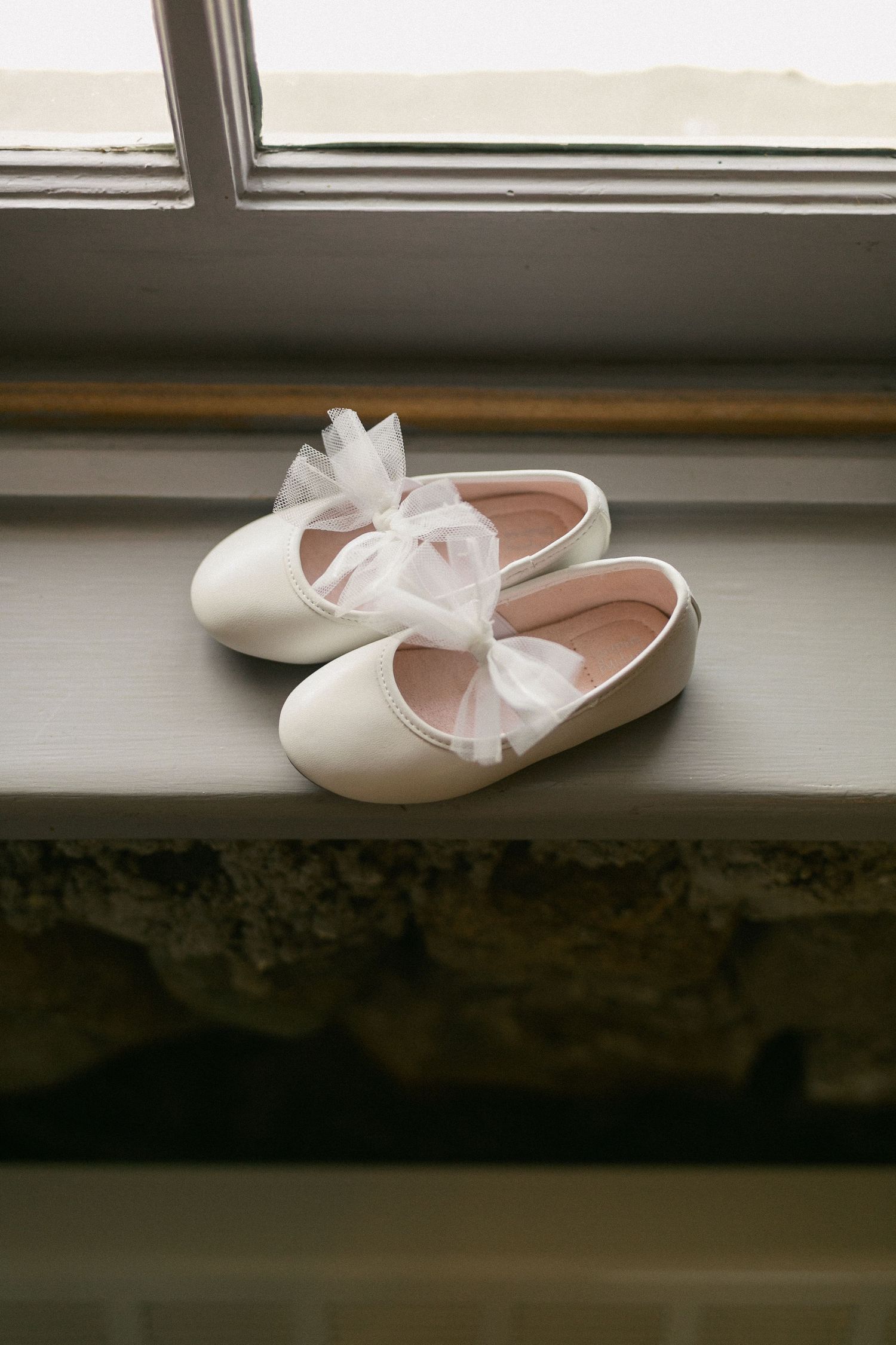 A pair of white leather baby shoes with delicate bows sits on a windowsill in soft natural light.