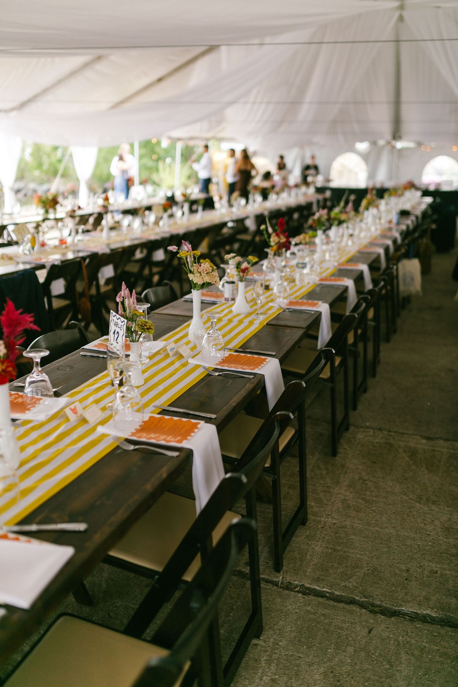 Long banquet tables set with white linens and yellow striped runners under a white tent for an elegant outdoor event.