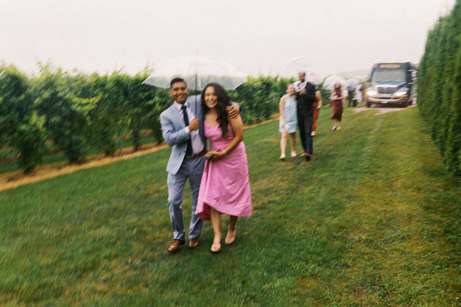 A couple and other wedding guests run through rain on a grassy hillside with scenic views in the background.