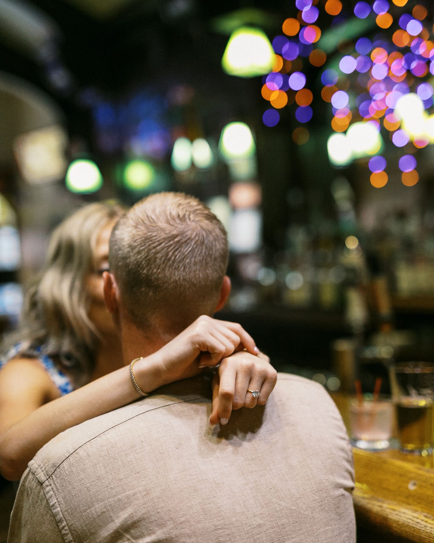 Couple sitting closely at a bar with colorful bokeh lights, woman resting hand on mans shoulder.