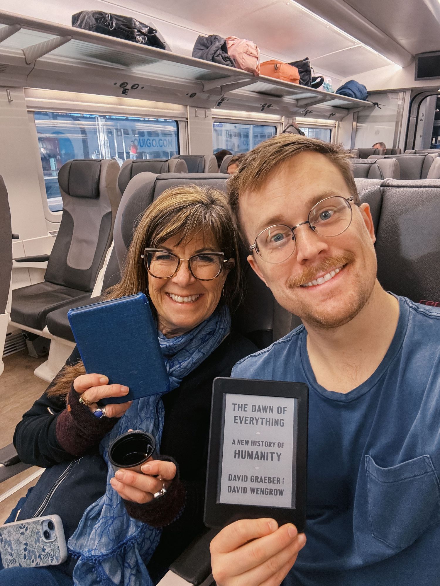 Two people on a train smiling holding books including The Dawn of Everything by David Graeber.