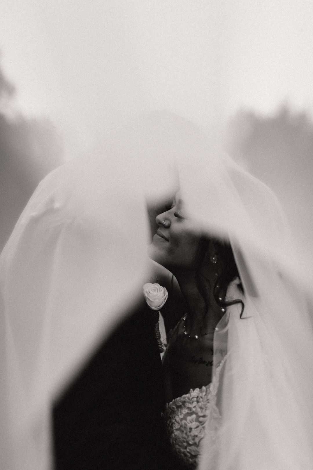 Romantic black and white photo of bride and groom kissing beneath flowing wedding veil outdoors.