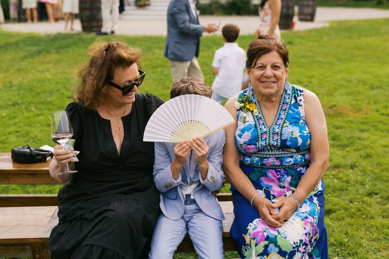 Three women sitting on a bench outdoors at a wedding, one holding a fan, another in a floral dress, smiling.